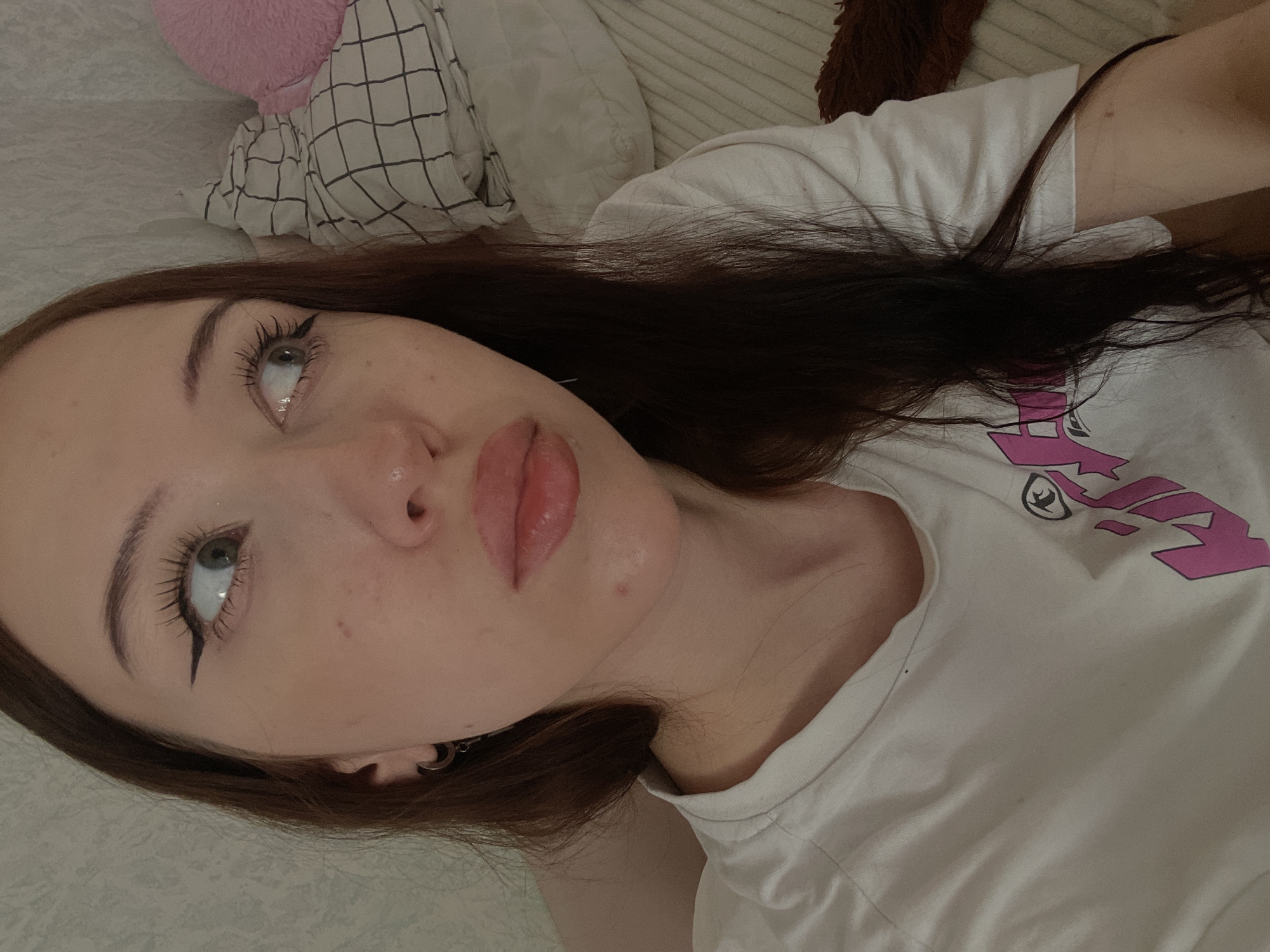 A young woman with long, dark hair is lying down, possibly in a bedroom. She is wearing a white t-shirt with a pink graphic on it. Her makeup includes winged eyeliner and mascara. She has a piercing in her left ear. In the background, there is a checkered pillow and a pink fuzzy object, likely a stuffed animal, on a bed or couch. The lighting suggests an indoor setting, potentially with natural light. The scene appears to be a casual, personal moment. The image does not provide any information about the city of Votkinsk, Russia.