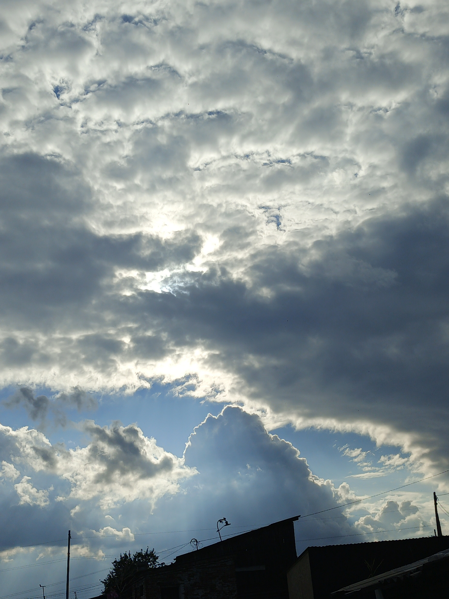 The image depicts a dramatic sky filled with varied cloud formations above silhouetted rooftops and a utility pole. In the distance, the city of Medvedyovka, Russia, is suggested by the presence of these elements. The sky is characterized by a mix of dark, heavy storm clouds and lighter, illuminated clouds, with rays of sunlight piercing through in places. The scene captures a moment of dynamic weather, with the sun partially obscured by the clouds, creating a high contrast effect. The foreground shows the dark outlines of buildings and a tall pole, implying a residential or urban setting. The overall impression is one of impending or passing weather, with the natural elements dominating the scene.