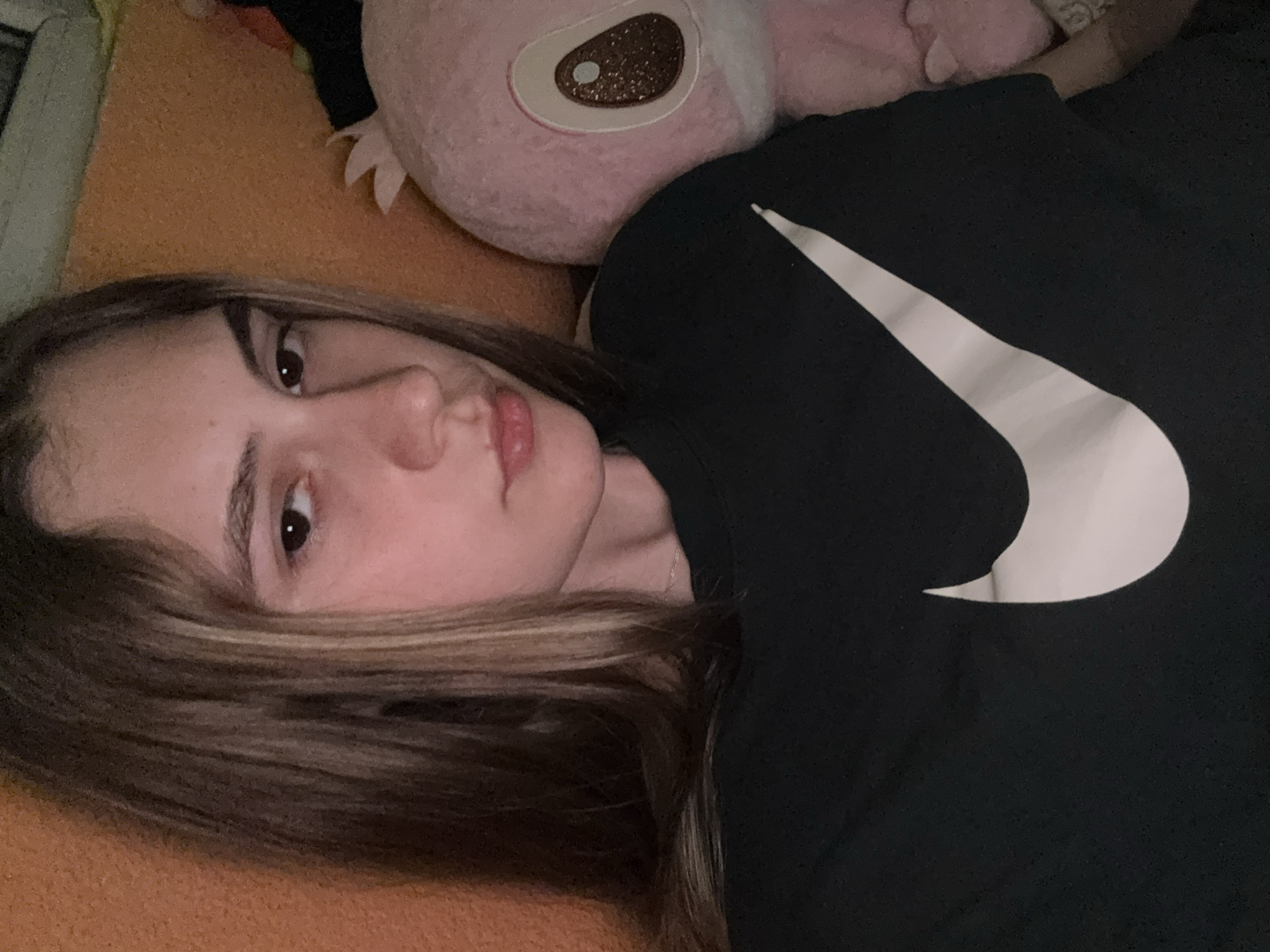 A young woman is depicted lying down indoors, in what appears to be a relaxed setting like a bed or couch, in Panagyurishte, Bulgaria. She is positioned on her right side, looking towards the viewer with a neutral expression. Her long, light brown hair, which has visible lighter streaks, is fanned out on an orange, textured surface that covers most of the background.
She is wearing a black t-shirt that features a large, white Nike swoosh logo prominently on the chest. Resting beside her head is a soft, pink plush toy. This toy has a distinctive large, dark eye with a brown, glittery center and a white reflective dot. A thin, silver necklace is visible on her neck. The lighting in the image is soft, suggesting an indoor environment with ambient or artificial light, illuminating her face clearly. The focus is primarily on her upper body and face, with the plush toy and Nike logo being significant elements of the composition.