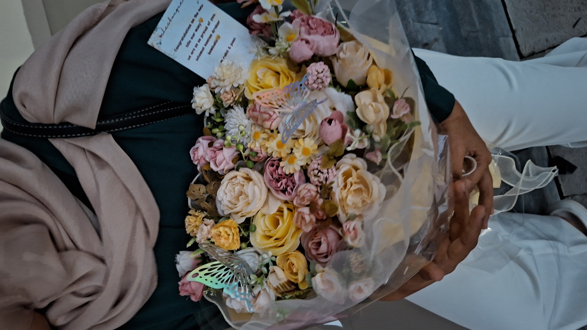 An individual wearing a dark green top and a beige hijab holds a bouquet of artificial flowers. The bouquet is wrapped in transparent cellophane and decorated with several light blue butterfly ornaments. A small white card with text written in blue is tucked into the flowers. The individual is also wearing white pants. The background shows a textured gray surface, possibly a wall or pavement, suggesting an outdoor urban setting. The presence of a card with handwritten text indicates a gift or congratulatory message. The overall scene suggests a personal occasion or celebration. Given the visual cues, the image may have been taken in Lhokseumawe, Indonesia, a city known for its significant Muslim population where hijabs are commonly worn.