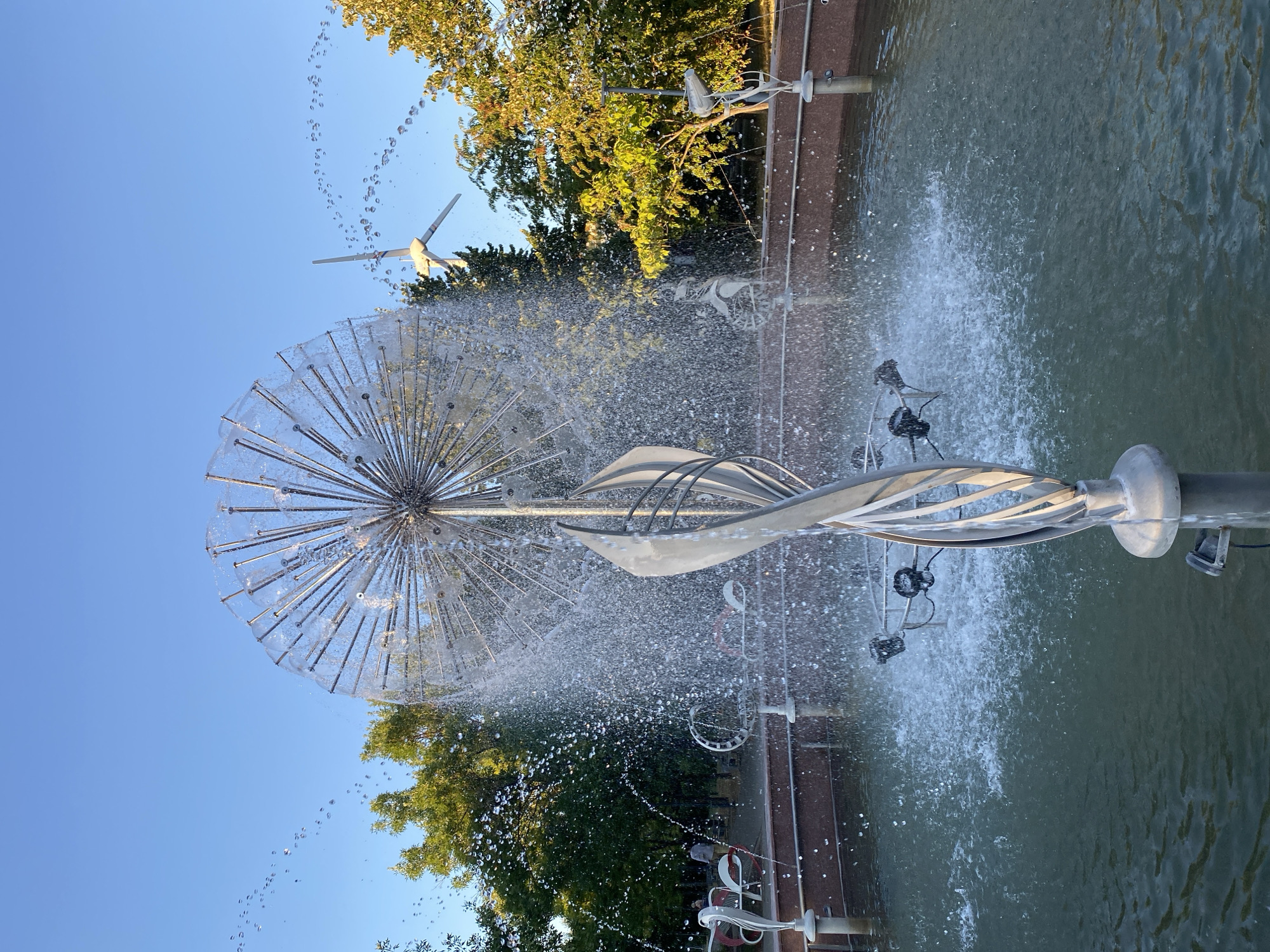 This image captures a modern art fountain in a public park, likely in Chișinău, Moldova. The fountain features a spherical metallic sculpture resembling a dandelion seed head, with numerous thin rods radiating outwards. Water sprays from the top of this sculpture, creating a shimmering cascade. Below the sphere, a twisted metallic element adds to the artistic design.
The fountain is situated within a circular basin with a red-brown rim. The water within the basin appears somewhat murky green. In the background, mature trees with green and yellow foliage line the park. The sky is a clear, bright blue.
Several other metallic sculptures are visible around the fountain's basin, some appearing abstract and others resembling stylized birds or seating. In the far background, a small wind turbine is visible, suggesting a modern or eco-conscious design for the park. A few people are casually strolling or sitting in the park area behind the fountain, indicating it is a public recreational space. The lighting suggests it is late afternoon or early evening, with the sun casting a warm glow on the scene.