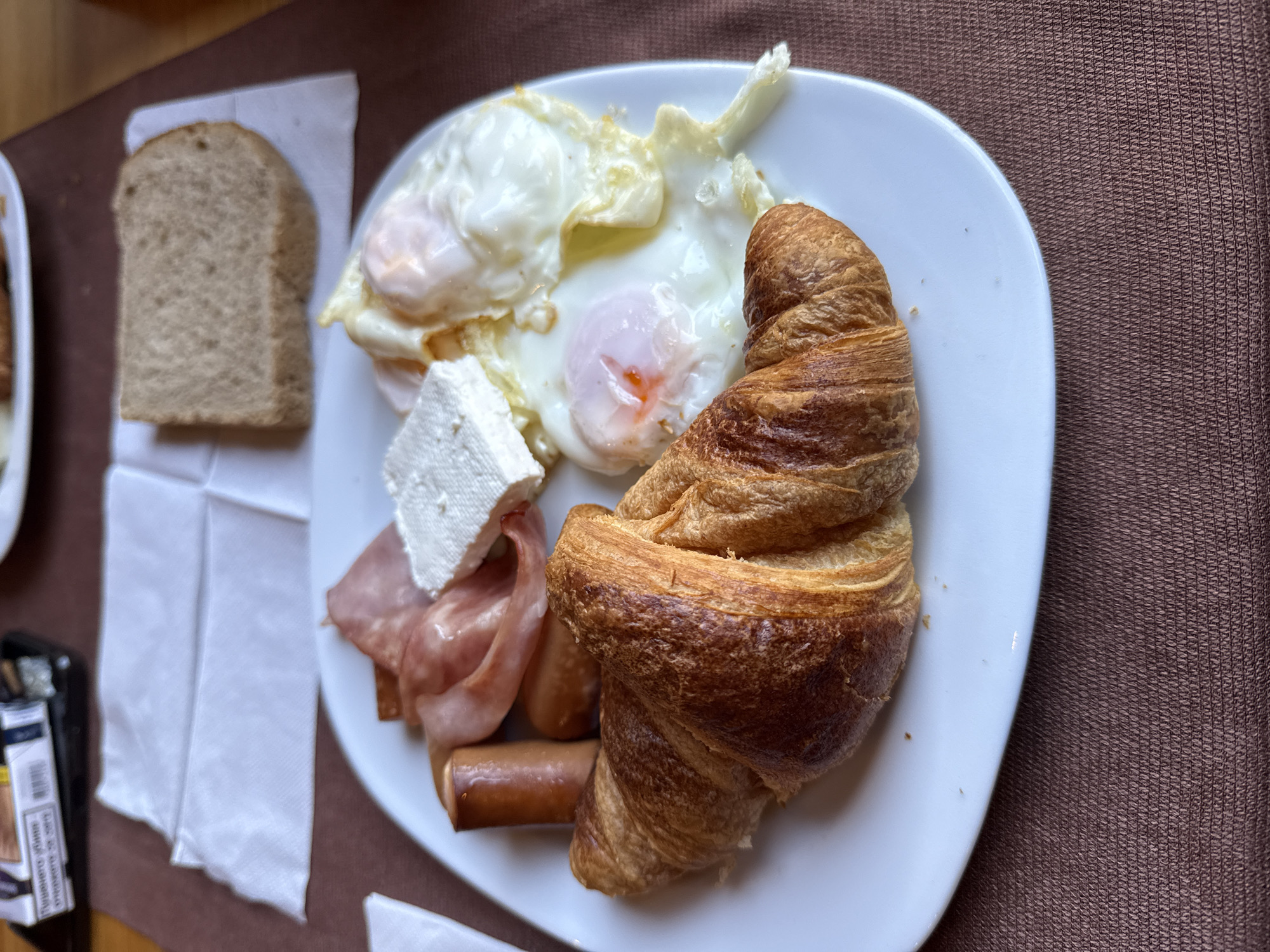 A plate of food is centered in the image, featuring two fried eggs, a croissant, a block of white cheese, two sausages, and sliced ham. A slice of bread is on a white napkin to the left. The plate rests on a brown surface. A cigarette pack is slightly visible in the bottom left corner. The location is Veliko Tarnovo, Bulgaria.