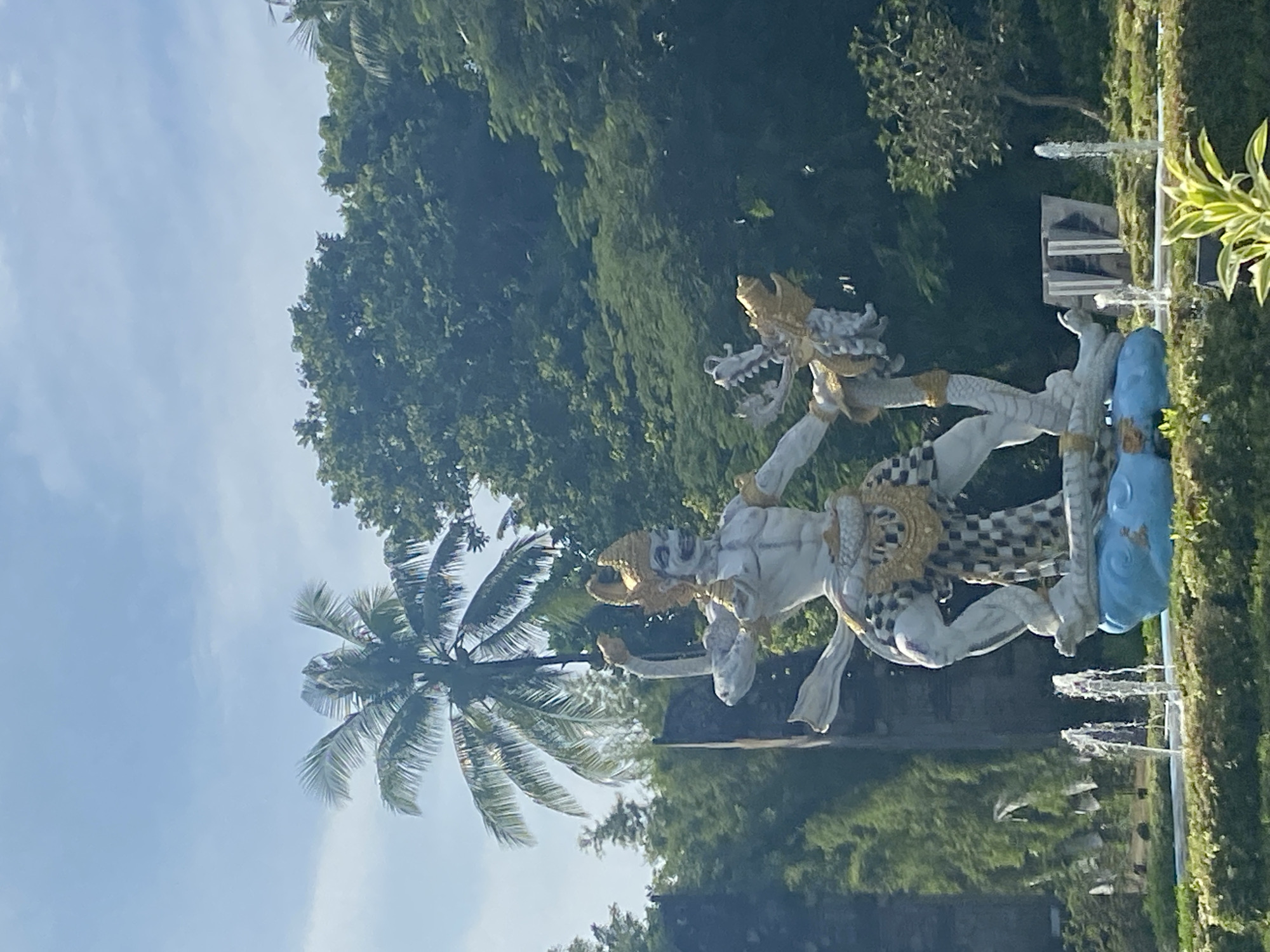 The image shows a statue of two figures intertwined in what appears to be a struggle or dance. The figures are primarily white with gold accents and checkerboard patterns on parts of their bodies. The upper figure has a dragon-like head. The statue is positioned next to a row of water fountains and lush green hedges. In the background, there are trees and a palm tree against a blue sky. The location is likely in Nusa Dua, Indonesia, given the tropical environment and Balinese-style statues often found in the area.