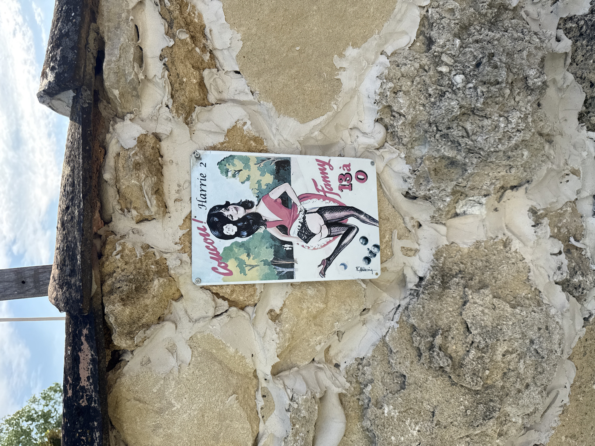 A sign depicting a cartoonish drawing of a woman with dark hair and a pink dress is affixed to a stone wall. The woman is shown in a provocative pose, with her legs spread and wearing stockings and garters. She is holding a set of balls, resembling those used in the game of pétanque. The sign has text that reads "Coucou! Harrie 2 Fanny 13à 0". The stone wall appears weathered and is composed of large, irregular stones. The background shows a cloudy sky and some greenery. The sign is slightly tilted, suggesting it may have been damaged or is not securely fastened. The overall impression is that this is a quirky or humorous sign placed in an outdoor setting. The sign's content suggests a playful or perhaps slightly risqué tone.