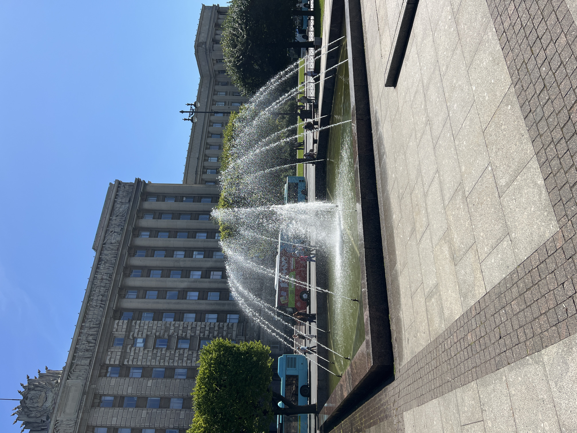 The image shows a public square in Saint Petersburg, Russia, featuring a large fountain in the foreground. The fountain has multiple jets spraying water upwards and outwards, creating a shimmering effect in the sunlight. Behind the fountain, a wide paved area leads to a street with two buses, one light blue and one red, and several people walking. Towering over the scene is a grand, imposing building with classical architecture, characterized by rows of windows, columns, and ornate decorative elements along its facade. The sky is clear and blue, indicating a sunny day. Trees line the street and part of the square, adding a touch of green to the urban landscape. The overall impression is one of a bustling city square on a pleasant day, with the fountain serving as a focal point.