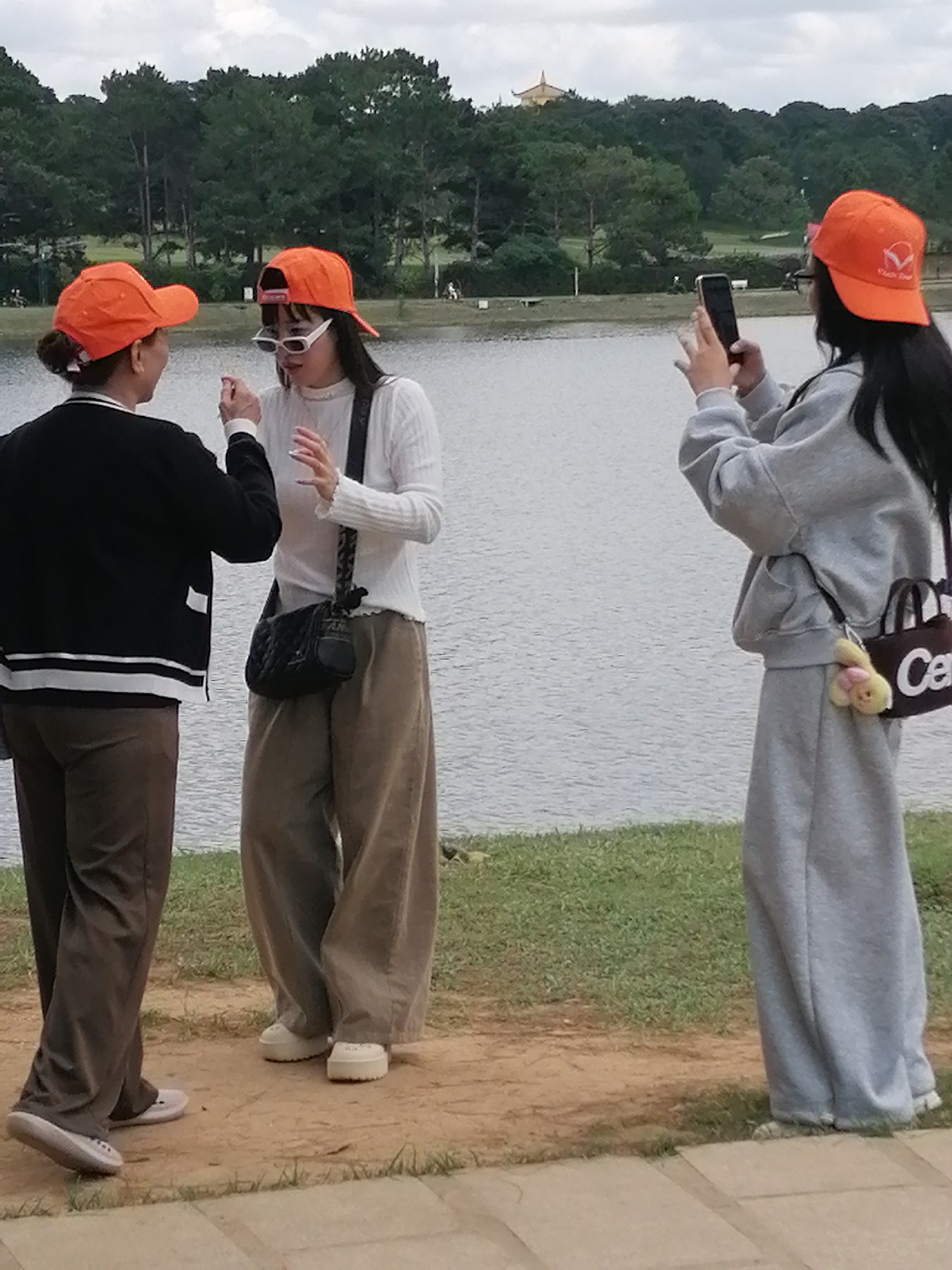 Three individuals are in the foreground, standing near the edge of a body of water, likely Xuan Huong Lake in Da Lat, Vietnam, given the surrounding pine forests. The sky is overcast.
On the left, a person wearing an orange baseball cap, black cardigan with white trim, and brown pants faces right. They are interacting with another individual.
In the center, a person also wearing an orange baseball cap and white sunglasses, is dressed in a white long-sleeved shirt and wide-legged brown pants. They have a black quilted crossbody bag and are gesturing with their hands while facing the person on the left.
On the right, a third person, wearing an orange baseball cap and a grey tracksuit, is facing away from the viewer and towards the water, holding a smartphone up, possibly taking a photo or video. They have a small brown bag with white lettering and a keychain attached.
The environment consists of a grassy bank leading down to the water, with a paved pathway visible in the bottom right corner. The background features a dense line of pine trees and a distant pagoda-like structure on a hill.