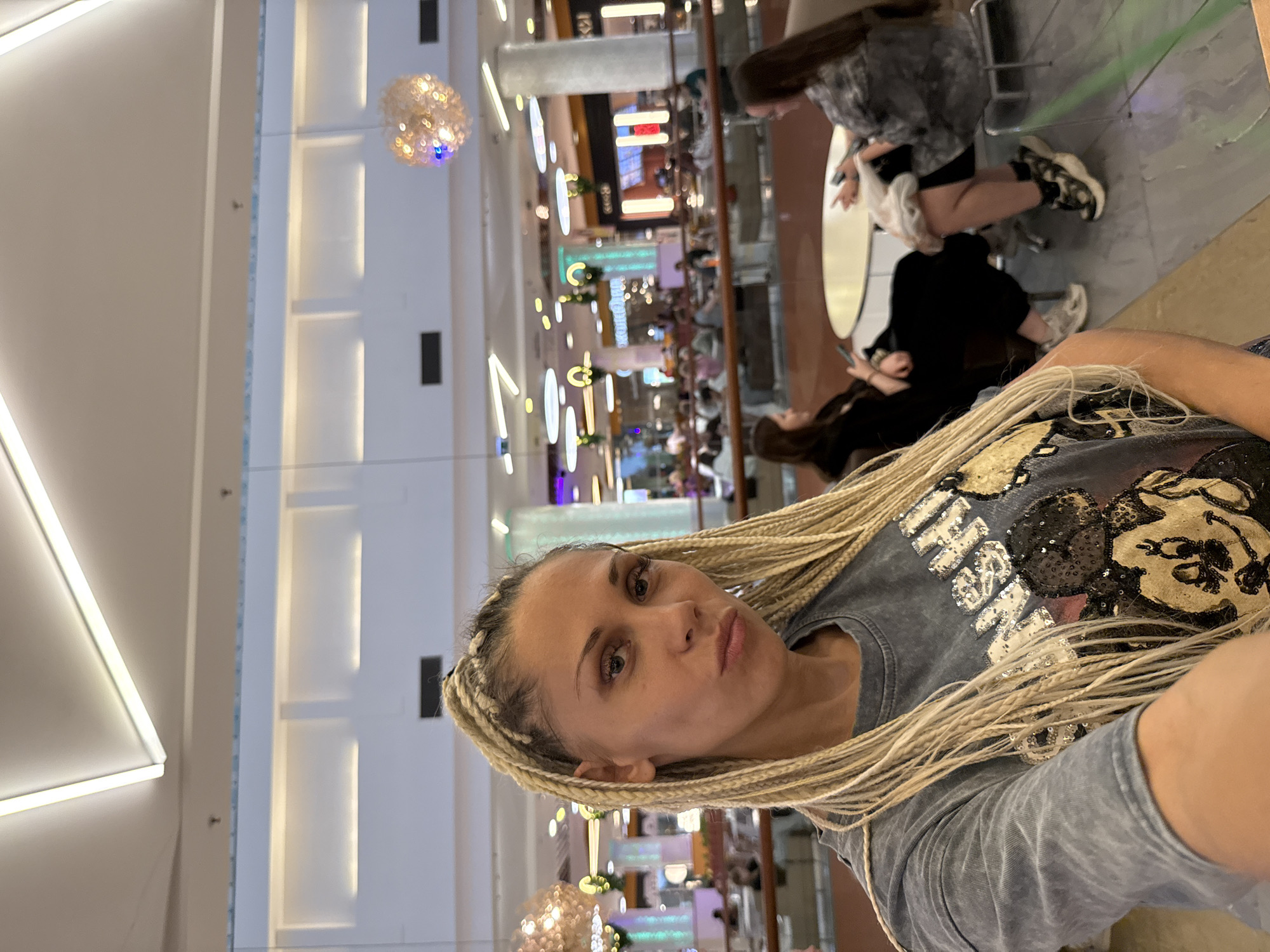 A medium shot captures a woman with blonde braids from the chest up, taken indoors in what appears to be a modern shopping mall or a large, well-lit public space with a high ceiling and geometric lighting fixtures. She is facing the camera with a pouty expression. She is wearing a gray t-shirt with a sequined graphic of Mickey Mouse and what appears to be a word that starts with "M". In the background, several people are seated at tables, some looking at their phones, while others are engaged in conversation. The environment is clean and contemporary, with polished floors and a sense of bustling activity. The location is Yaroslavl, Russia.