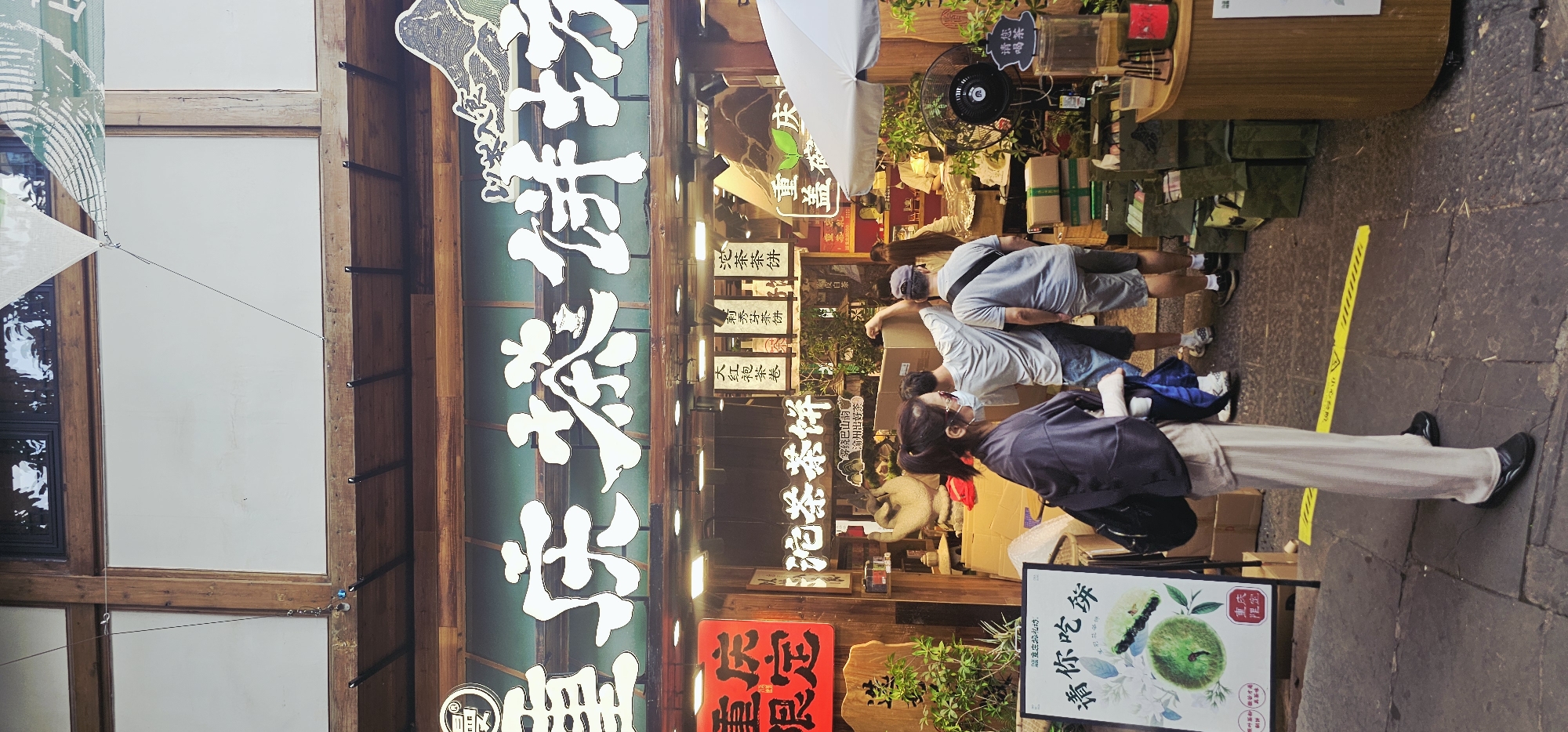 The image shows the exterior of a tea shop, possibly named "Chongqing Tea Cake Shop", located in Yuzhong District, China. The shop facade features large white Chinese characters above the entrance, with smaller signs and hanging banners visible. The shop displays various tea products, boxes, and green plants. Several individuals are present outside the shop. A woman in a dark purple garment and beige pants stands in the foreground on the left, facing away from the camera, wearing glasses and a mask. To her right, three men are entering or exiting the shop. One man in a light grey t-shirt and dark shorts is carrying a large cardboard box. Another man in a grey t-shirt and grey shorts is walking behind him. A third man in a light blue t-shirt and denim shorts is also visible. The ground is paved with cobblestones. A sign in front of the woman on the left displays an image of a green fruit or pastry and Chinese text. A fan and a display counter are visible to the right of the entrance.