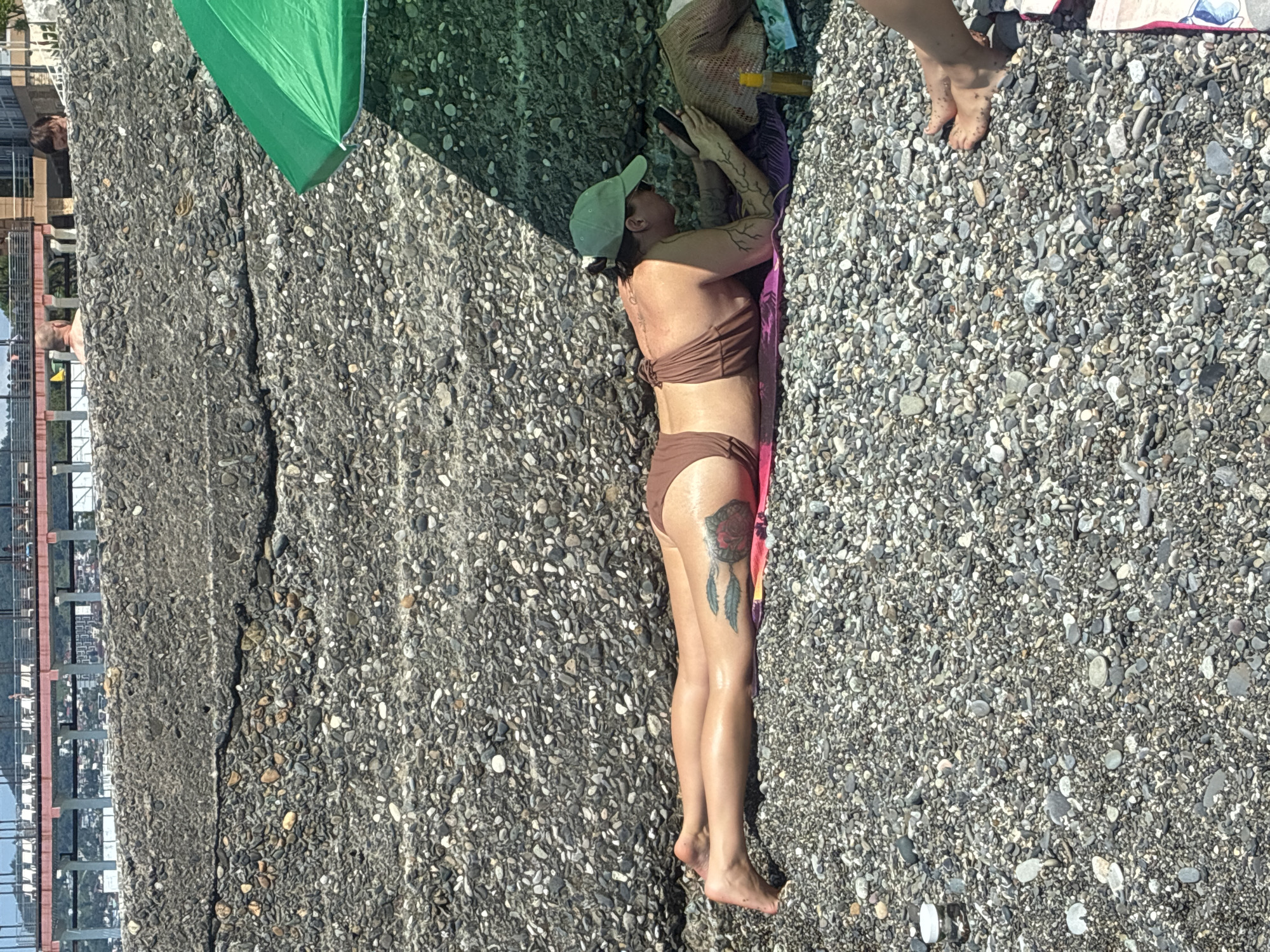 A woman is lying prone on a pebbly beach in Sochi, Russia, facing a phone she holds in her hands. She wears a brown bikini and a green baseball cap. A large green umbrella shades her from above, casting a shadow on the ground. Her legs are extended, and a tattoo of a rose and feathers is visible on her left thigh. A bottle of sunscreen sits on the towel beside her. In the background, a concrete embankment and a multi-story building complex are visible.