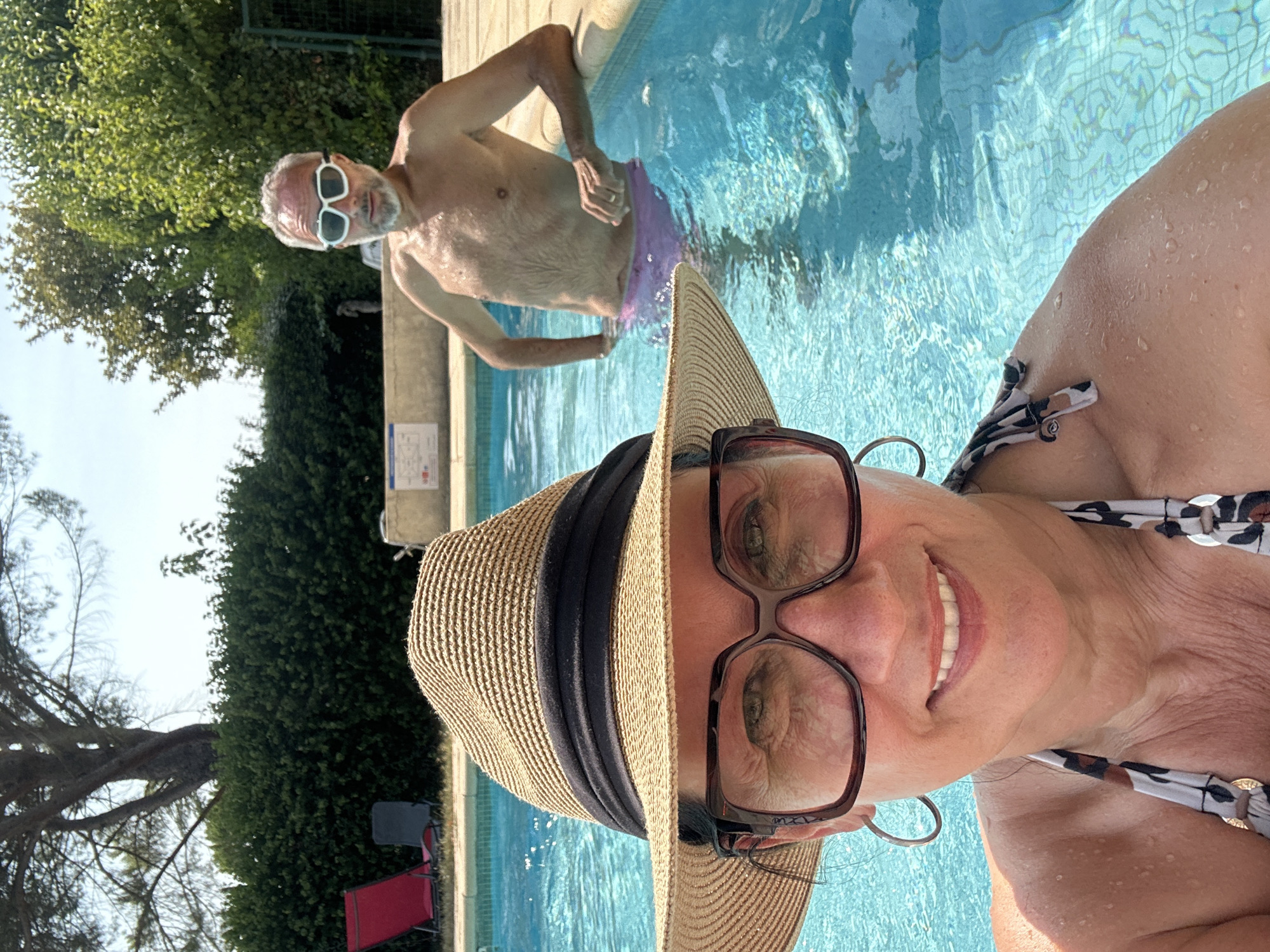 The image shows a couple in a swimming pool on a sunny day. The woman is in the foreground, wearing a straw hat and glasses, smiling at the camera. She is wearing a patterned bikini. The man is in the background, wearing goggles and purple swim trunks. He is leaning on the edge of the pool. The pool is surrounded by lush green trees and a hedge. The scene appears to be in a backyard or resort setting. The specific location within Saint-Marcel-lès-Sauzet, France, cannot be determined from the image alone.