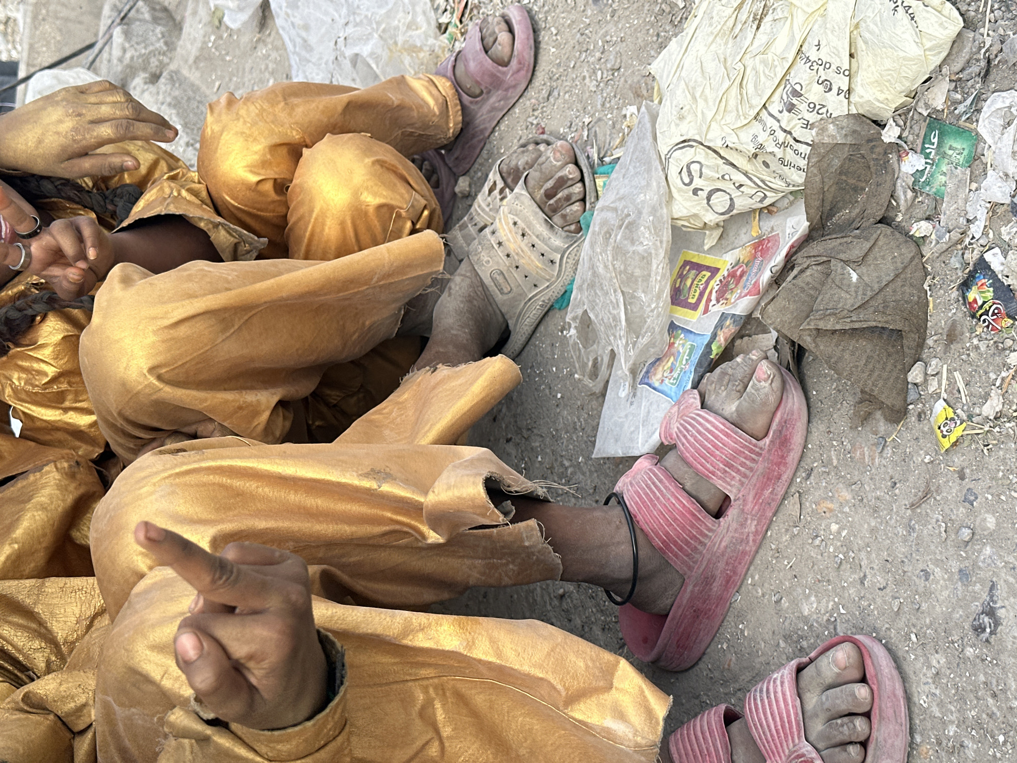 The image depicts two individuals adorned in golden clothing, lying amidst litter on the ground in what appears to be an outdoor setting. The location is identified as Quetta City Tehsil, Pakistan.
One individual is wearing pink sandals and has a black anklet. The other individual wears white sandals with star cut-outs, and a purple sandal lies nearby. The feet of both individuals appear dusty and dirty.
The surrounding litter includes crumpled plastic bags, food wrappers, and other discarded items, suggesting an area with improper waste disposal. The image's composition is from an elevated viewpoint, looking down at the scene. One individual is gesturing with their hand.