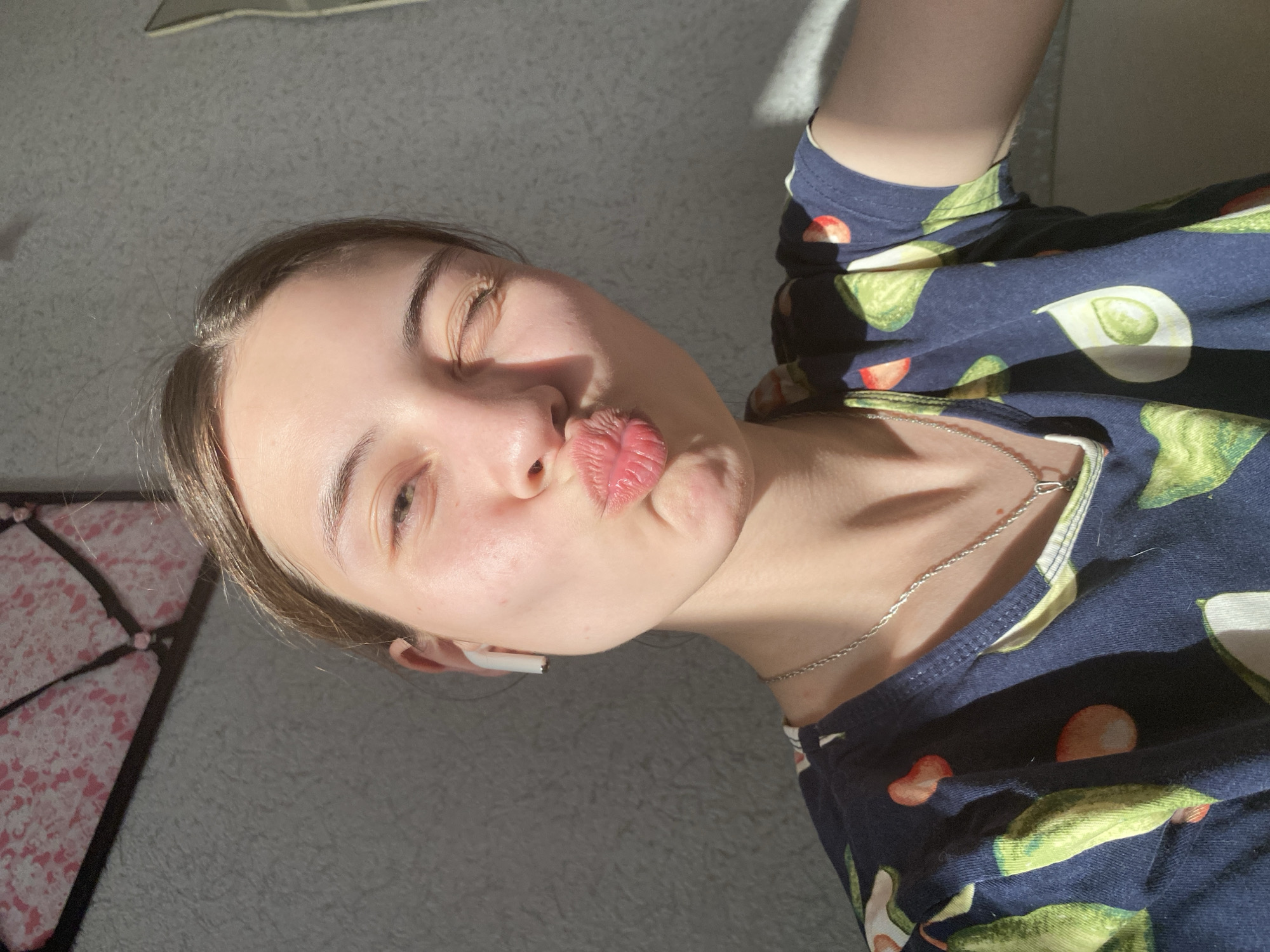 A young woman with light skin and brown hair is posing for a close-up, selfie-style photograph. She is pursing her lips in a kissing gesture and glancing to her left. She is wearing a navy blue t-shirt with an avocado print. A silver chain necklace is visible around her neck. A white wireless earbud is in her left ear. The background includes a gray carpeted floor, a patterned object (possibly a rug or pillow) in the lower left corner, and diffused sunlight casting shadows across the scene. There is no information provided to suggest a location in the city of Votkinsk, Russia.