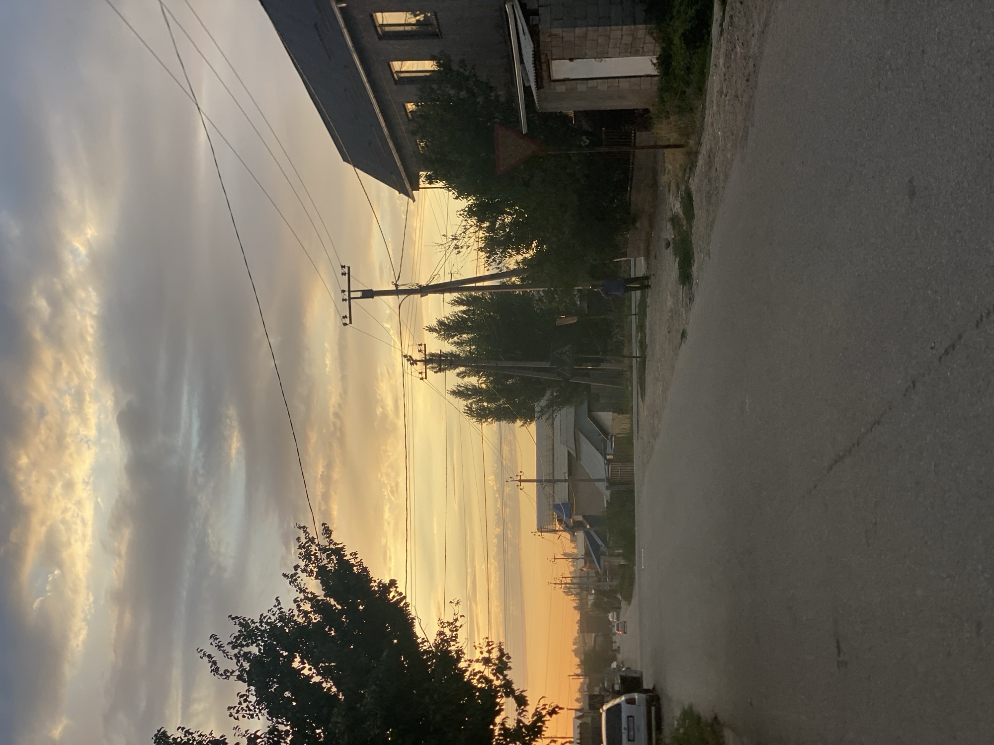 The image captures a street scene at dusk in Bishkek, Kyrgyzstan. The sky is filled with dramatic clouds, illuminated by the warm hues of sunset, ranging from orange to yellow and gray. A paved road runs through the center of the frame, with a few cars visible, including a white car parked on the left side and other vehicles driving in the distance. On the right side of the road, a dark, two-story building with windows reflecting the sky is prominent. Adjacent to it is a smaller, single-story structure made of brick with a white door. A triangular yield sign stands near the road. Trees, some with dark foliage, line the street, and utility poles with power lines are scattered throughout the scene, indicating the presence of infrastructure. A lone figure in dark clothing is standing on the side of the road, facing away from the camera, near the yield sign. The overall impression is a quiet, residential street at the end of the day.