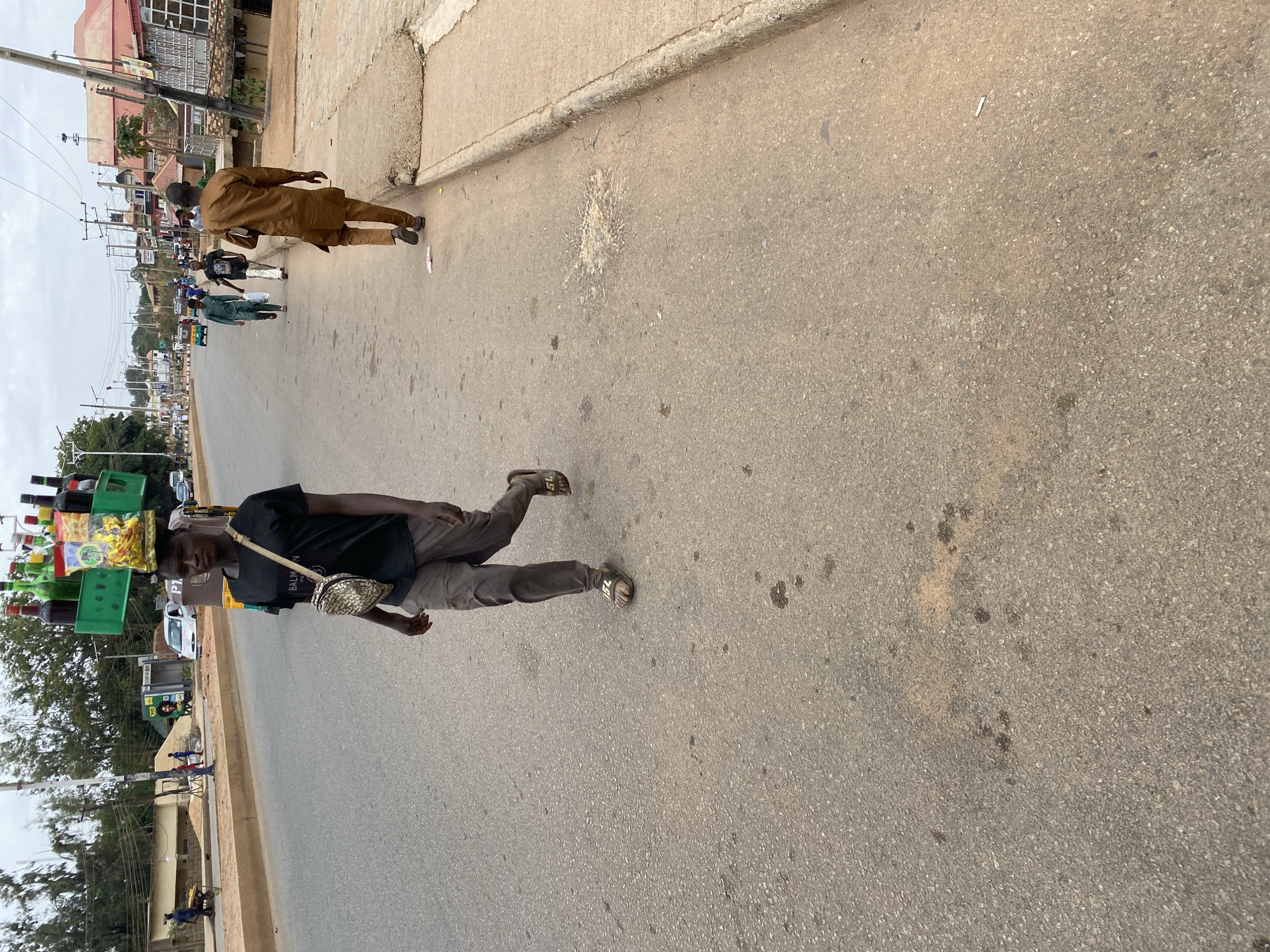 The image, taken in Bukuru, Nigeria, shows a street scene. A man in a black shirt and brown pants is walking on the road, carrying a green container filled with bottles and other items on his head. He is also wearing a small bag across his shoulder. Another man in a brown outfit is walking in the same direction in the distance. The road is asphalt, with a sidewalk on the right side of the frame. There are buildings, trees, and utility poles visible in the background, along with some other people and vehicles in the distance. The sky is overcast.
