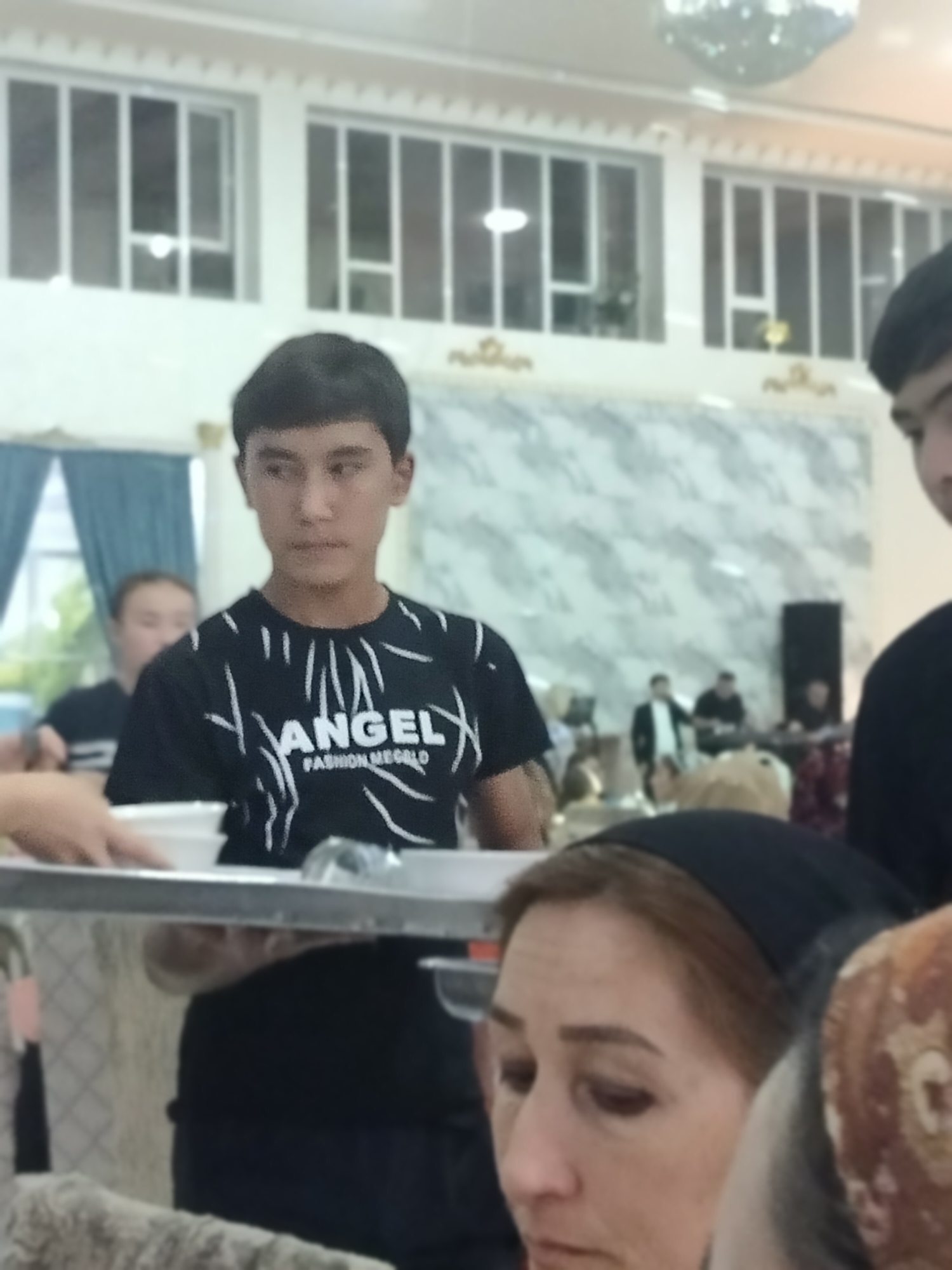 The image depicts an indoor scene, likely a gathering or event, with multiple individuals present. In the foreground, a young man is wearing a black t-shirt with white graphics that appear to say "ANGEL FASHION MEGALO." His gaze is directed slightly to the right. To his left, a person's hands are visible holding a white bowl. To the right and slightly behind the young man, a woman is wearing a black headscarf and is partially visible. In the background, several other individuals are present, some appearing to be musicians or performers on a stage. The environment includes a patterned wall, windows with blinds, and what looks like a disco ball suspended from the ceiling. The overall impression is one of a social gathering. Based on the provided crops and general visual cues, the location is identified as Ақсуабат, Kazakhstan.