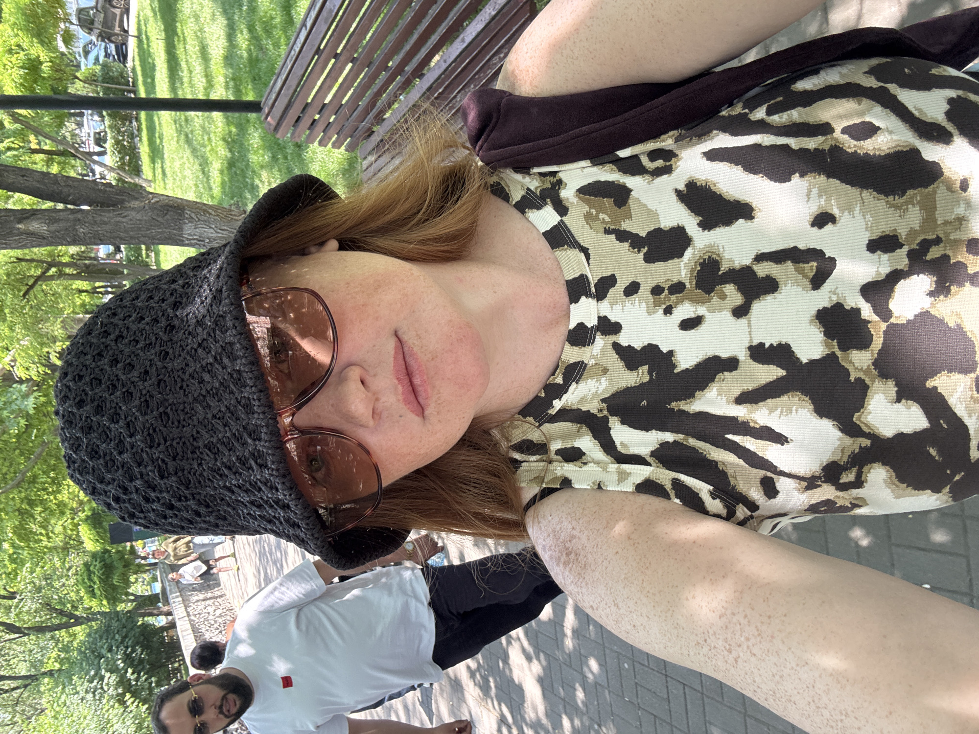 A woman with red hair, wearing a black crocheted bucket hat, sunglasses, and a leopard-print tank top, takes a selfie in a park. She has freckles on her face and arm. In the background, a man in a white t-shirt and dark pants walks on a paved pathway. Other people are visible in the distance, and there are trees, grass, and a park bench. The scene appears to be a sunny day in a park, possibly in Yerevan, Armenia, given the context.