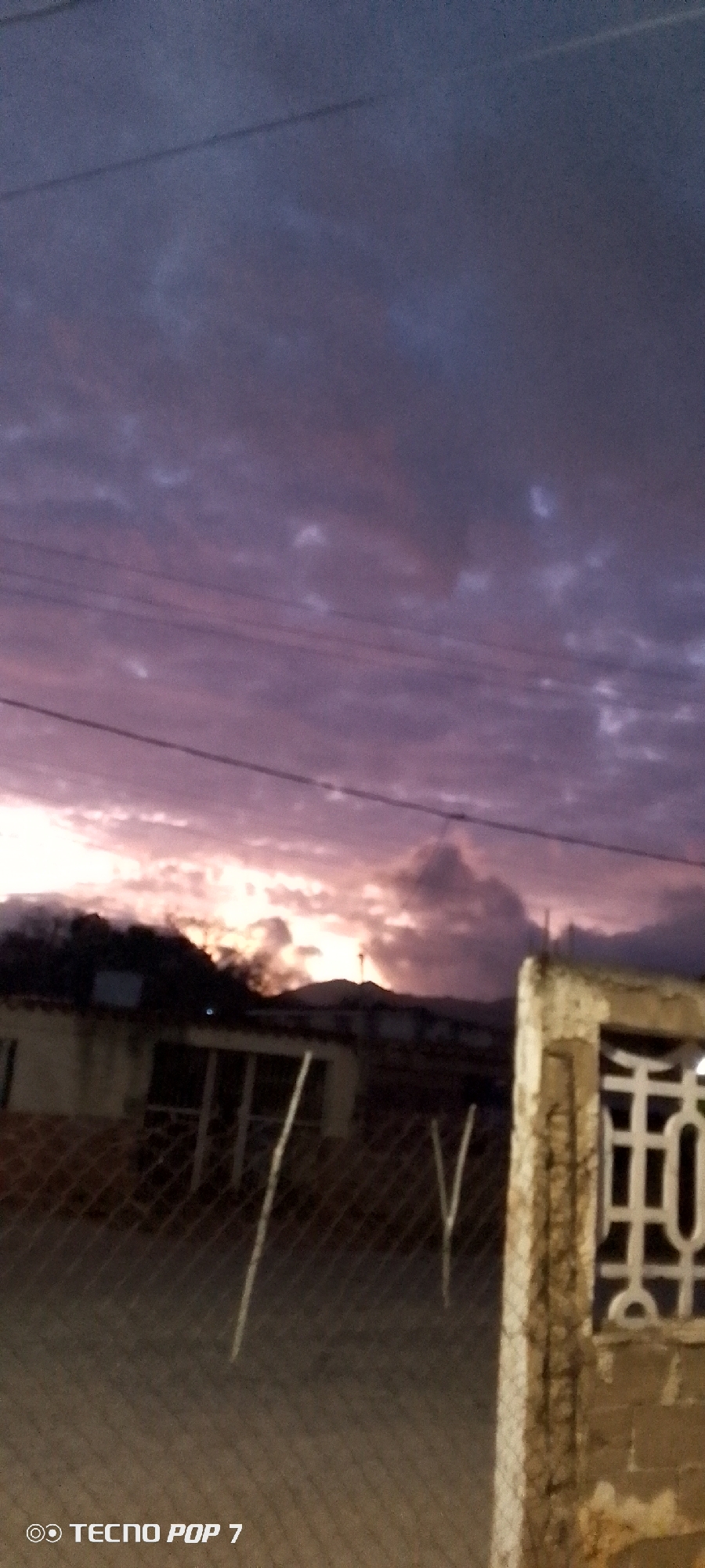 The image shows a twilight scene in Altagracia, Venezuela. The sky is filled with purplish-gray clouds, with a bright, light-colored area on the horizon indicating the setting sun. Power lines stretch across the upper part of the frame. In the foreground, a chain-link fence partially obscures the view of a building and yard. To the right of the fence is a concrete post with a decorative, geometric design. The lower left corner has the words "Tecno Pop 7." In the background, behind the building, are hints of mountains silhouetted against the fading light.