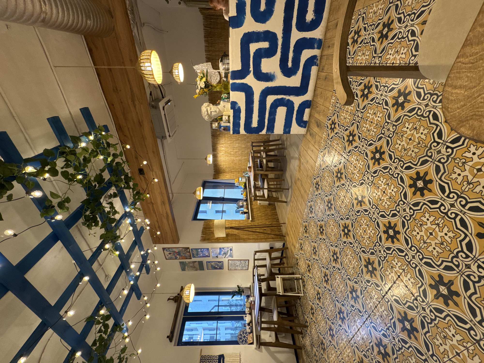 This is an interior shot of what appears to be a restaurant. The decor is eclectic, combining elements of modern and traditional styles. The flooring consists of patterned tiles in shades of yellow, brown, and dark blue, covering a large portion of the scene. Tables and chairs are arranged in rows, suggesting a seating layout for customers. There are two visible window views of Izhevsk, Russia.
Along one wall, there's a decorative blue lattice structure adorned with greenery and string lights, creating a whimsical atmosphere. Lighting fixtures hang from the ceiling, providing illumination. A counter or bar area is present, featuring a design with blue and white patterns. The setting is well-lit, suggesting an inviting and comfortable ambiance for dining.