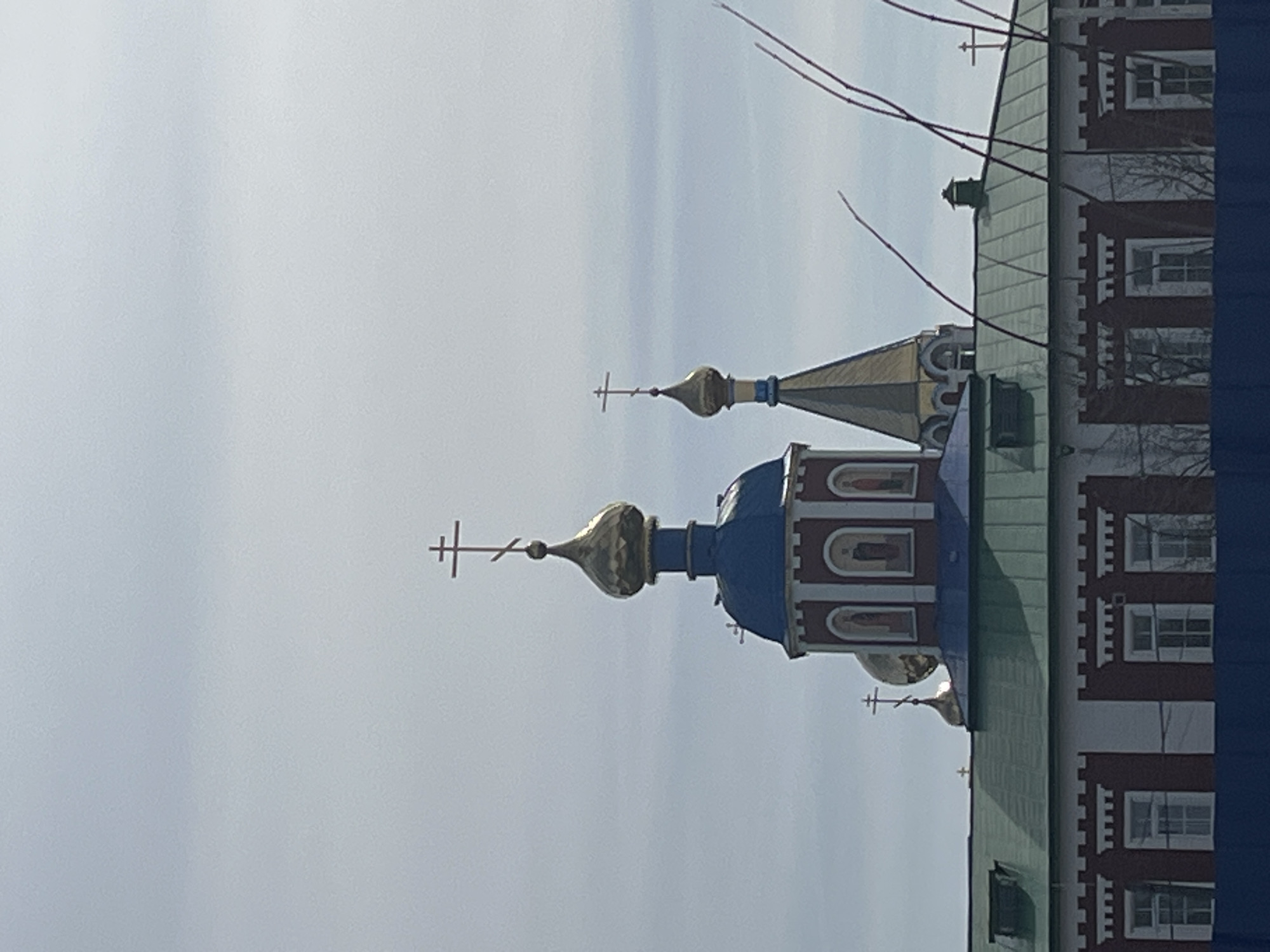 The image depicts the Church of St. Nicholas the Wonderworker in Nor'ya, Russia. Two onion domes with crosses are visible against a pale sky. The larger dome is blue with gold accents, while the smaller dome is primarily gold. A section of the church building is visible on the right side of the image, featuring multiple windows and a red and white facade. Branches of trees are visible in the upper right corner. The perspective is from a low angle, looking up at the church.