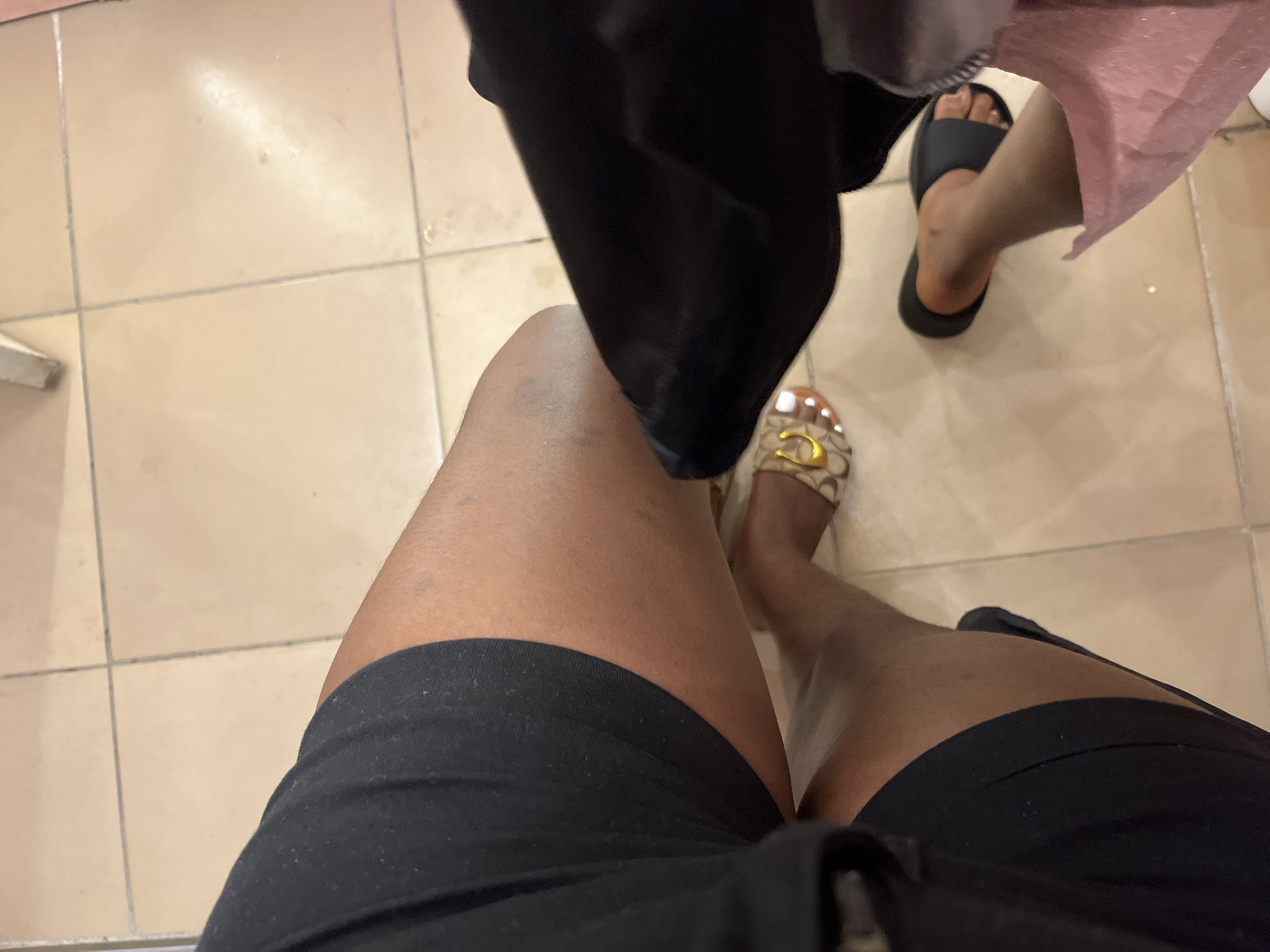 A low-angle, close-up shot shows the legs and feet of at least two individuals, seated on a light beige tiled floor. The person in the foreground on the left wears black shorts, with their right leg bent and foot bare. Their left leg is also bent, with a black shorts leg visible. To the right, another individual's legs are visible. One leg is bare and wearing a black slide sandal, while the other is covered by a pink fabric. The background consists of the tiled floor with visible grout lines. The lighting is somewhat dim and warm. There are no specific landmarks to identify a location in the city of Hawayi, Nigeria.