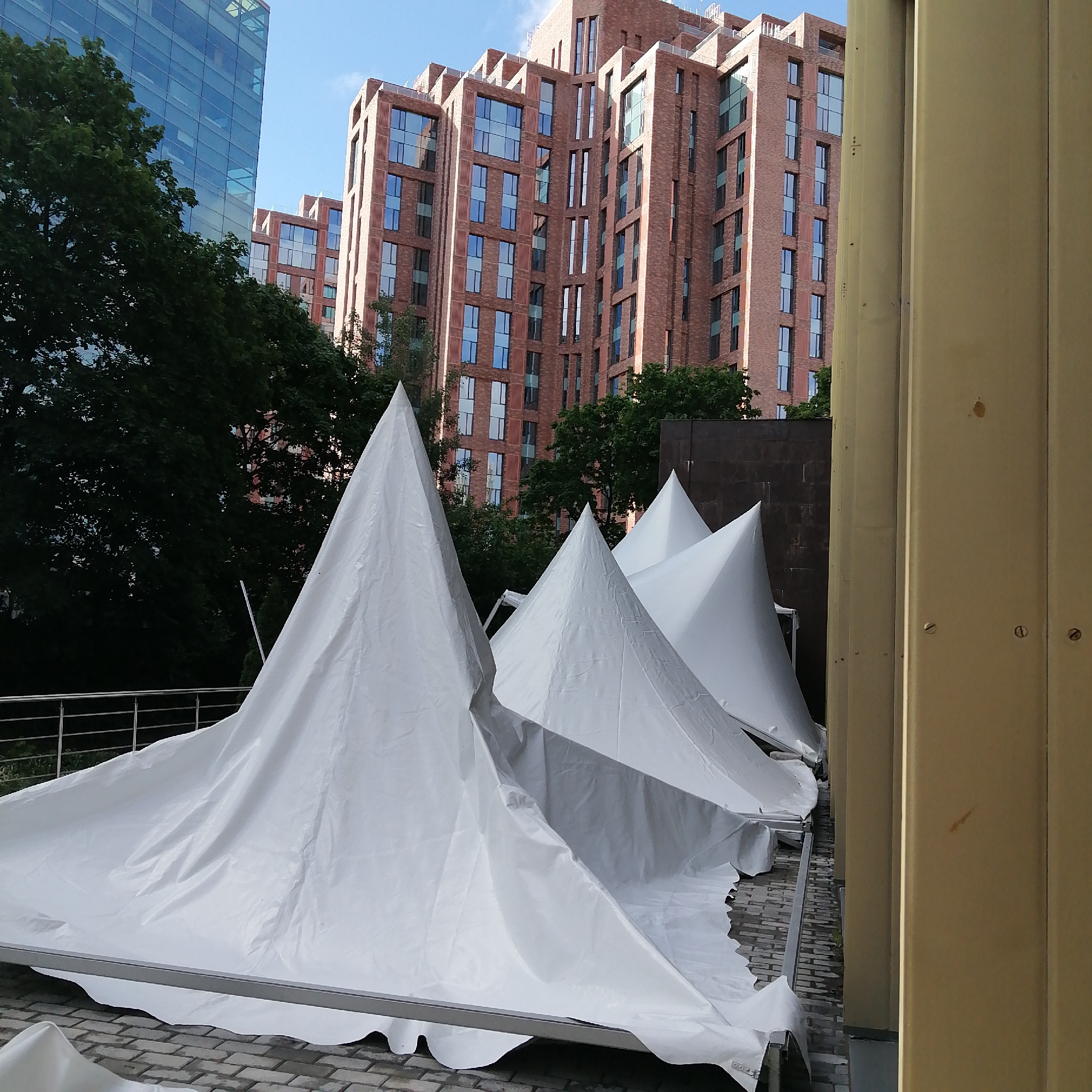 The image depicts several white conical tents erected on a paved surface, likely a public space in Moscow, Russia, given the architectural style of the surrounding buildings. The tents appear to be part of an event setup or temporary installation. Behind the tents, there are trees and two multi-story buildings. One is a modern glass and steel structure, while the other is constructed of reddish-brown brick with multiple windows, indicative of contemporary residential or commercial architecture. The light suggests it is daytime, and the scene is outdoors. There are no people visible in the frame.