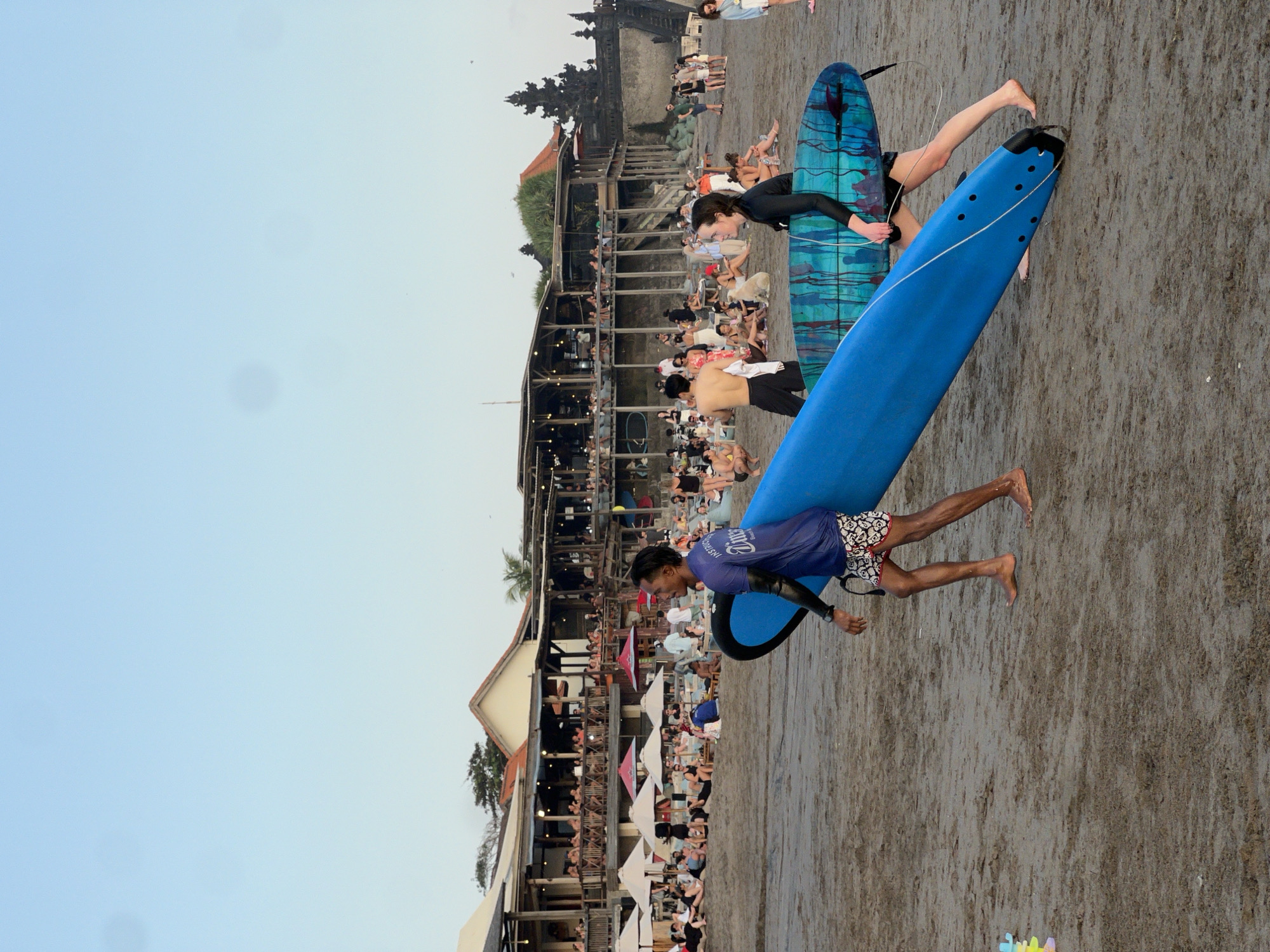 In the city of North Kuta, Indonesia, on a sandy beach, two individuals are carrying surfboards. The person on the left, a male, is wearing a wet blue t-shirt and patterned shorts. The person on the right, a female, is wearing a black wetsuit. They are walking from right to left across the frame. Behind them, a beach area is populated with numerous people sitting and standing, many under umbrellas. Several open-air buildings with tiered seating are visible in the background, suggesting a beachfront dining or recreational area. The sky is a clear light blue, indicating daytime.