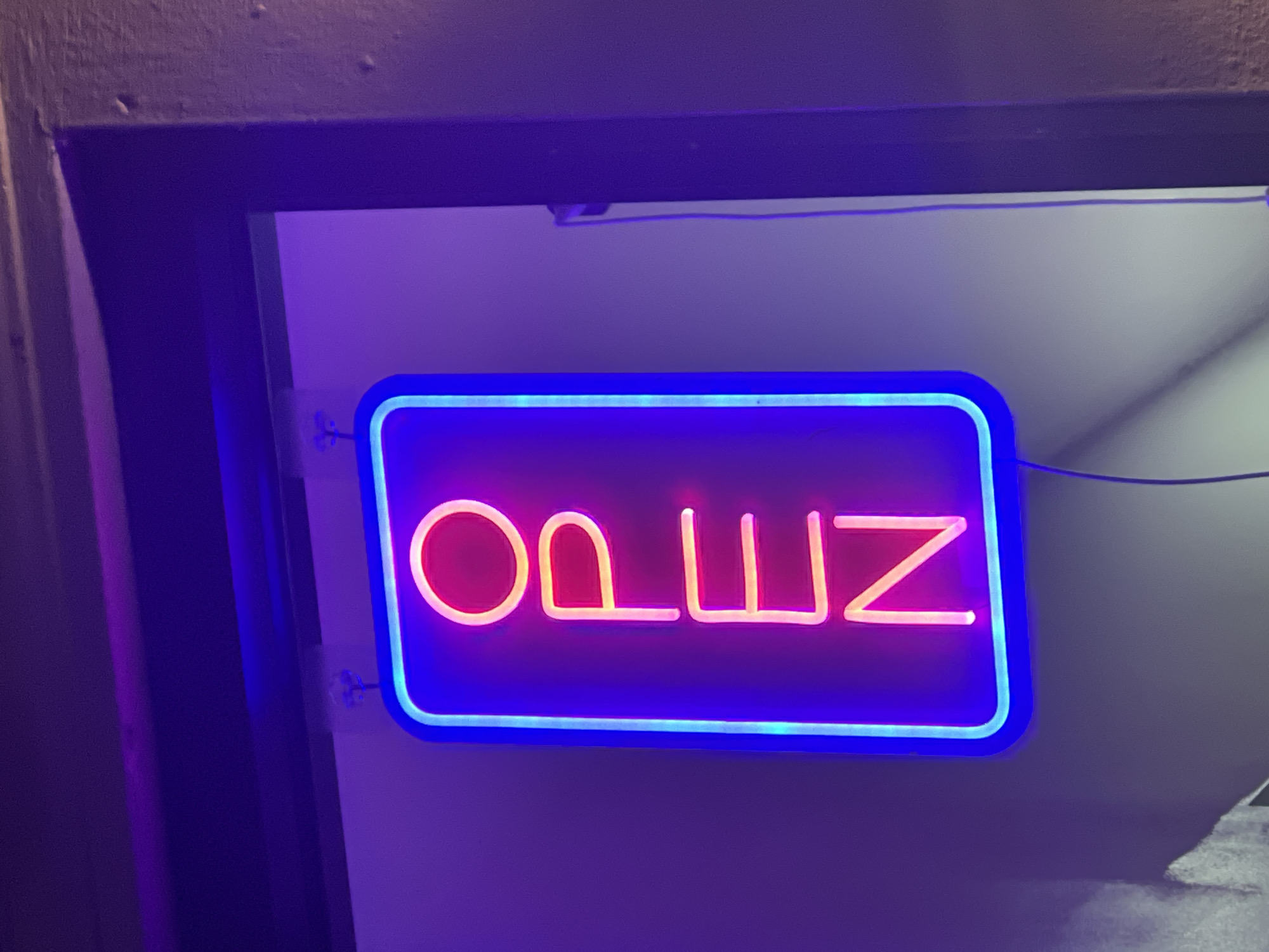 The image shows a neon sign that reads "OPEN" in reverse. The letters are rendered in a pink neon tube, set against a dark purple background. The entire sign is framed by a border of blue neon tubing. The sign appears to be mounted within a doorway or archway, possibly leading into a shop or business. The surrounding architectural elements are dimly lit and indistinct. The sign seems to be in London, United Kingdom.