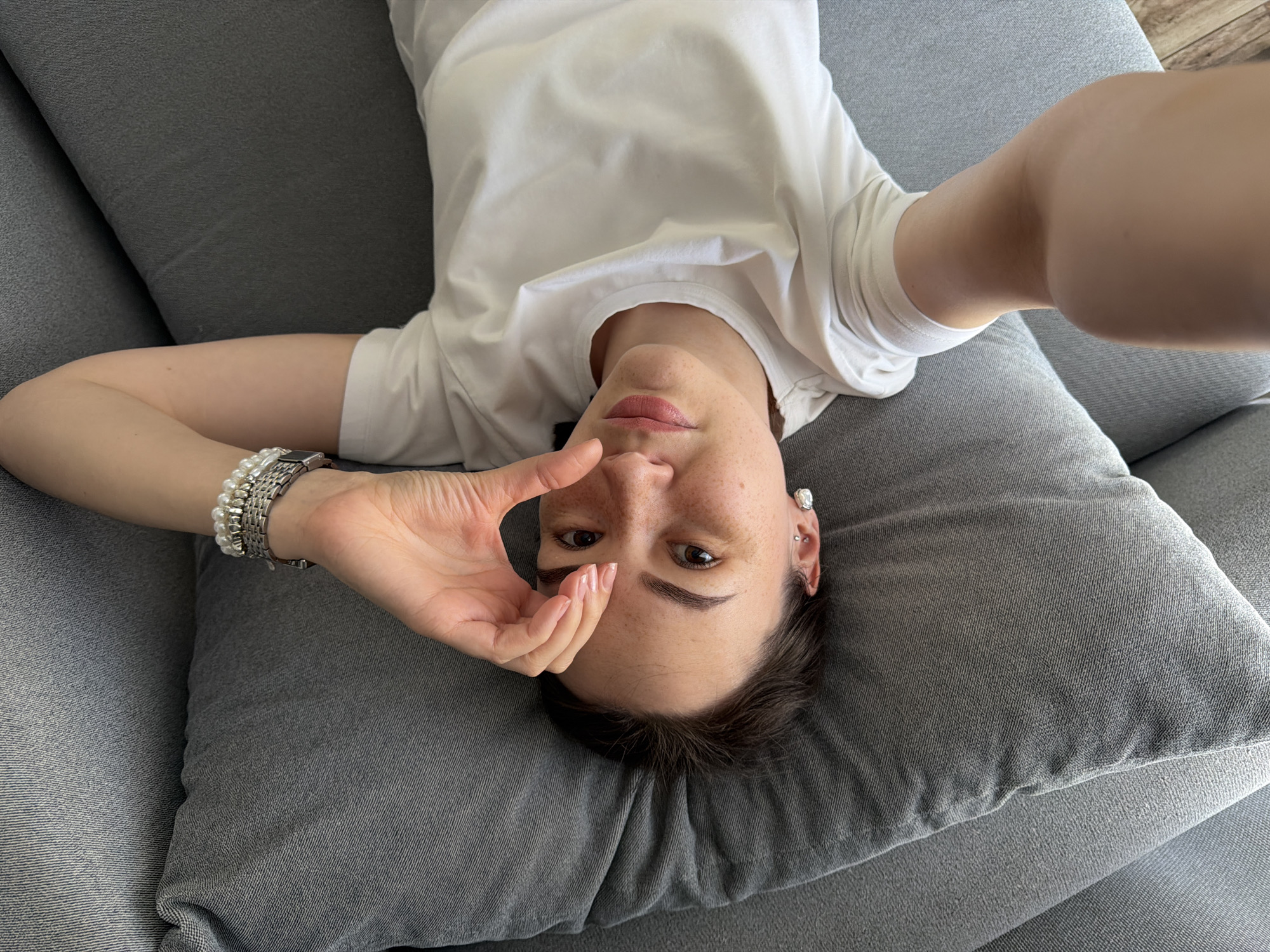 A woman is lying on a gray couch. She wears a white t-shirt and a bracelet. The location in the city of Chaikovsky, Russia is not discernable from the image.