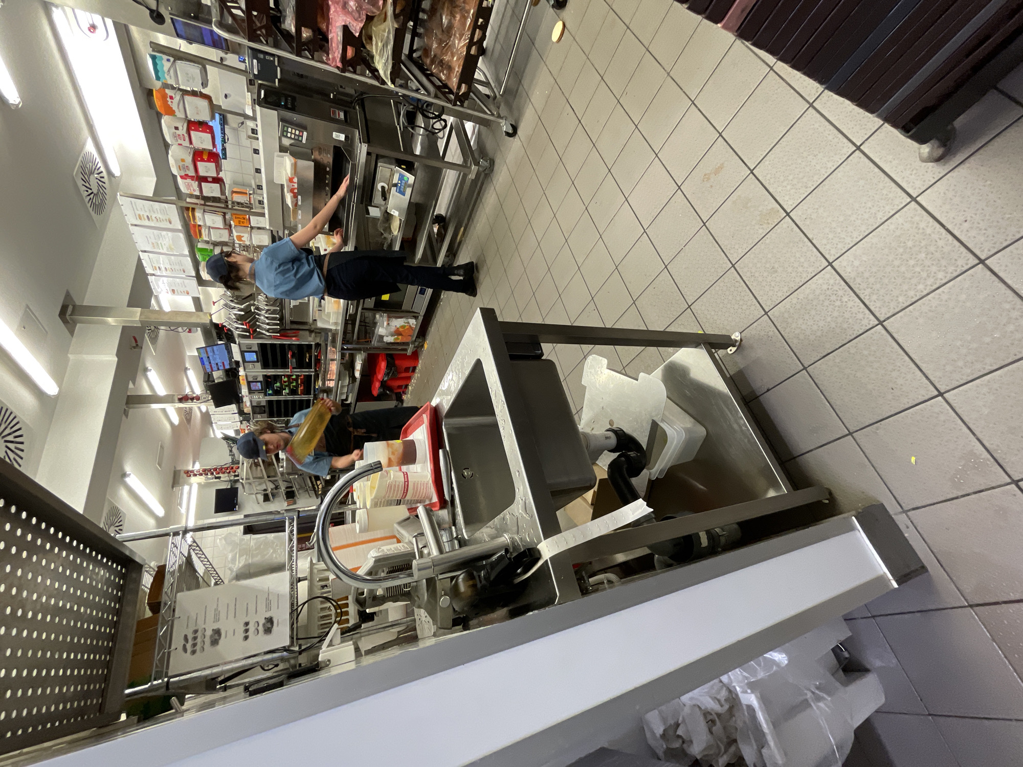 The image depicts the interior of a fast-food restaurant kitchen, likely in Boyarka, Ukraine. Two employees in blue uniforms and dark aprons are visible. One, wearing a blue cap, is standing near a sink area, holding a yellow container. The other employee, also wearing a blue cap, is positioned behind a stainless steel counter, interacting with a piece of kitchen equipment. The environment is characterized by stainless steel surfaces, tiled flooring, and various food preparation and serving items, including containers, trays, and dispensers. A sink with a faucet and a roll of paper are prominently featured in the foreground. The overall scene suggests an active work environment within a commercial kitchen setting.