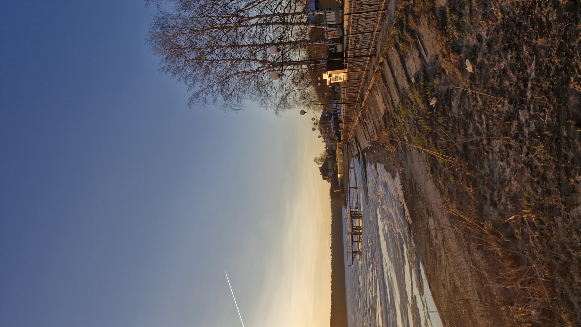 The image depicts a waterfront scene in Otchor, Russia. A body of water, possibly a lake or river, is visible in the foreground. A pier or dock extends into the water. On the bank, a concrete embankment is present. A wrought iron fence with globe-shaped light fixtures runs along the top of the embankment. Bare trees are located behind the fence. Several small buildings and vehicles can be seen in the background. The sky is predominantly blue, transitioning to a yellowish hue near the horizon, suggesting either sunrise or sunset. A contrail from a jet is visible in the sky. The overall light indicates that the scene is captured during the golden hour. The vegetation on the bank appears dry and dormant.
