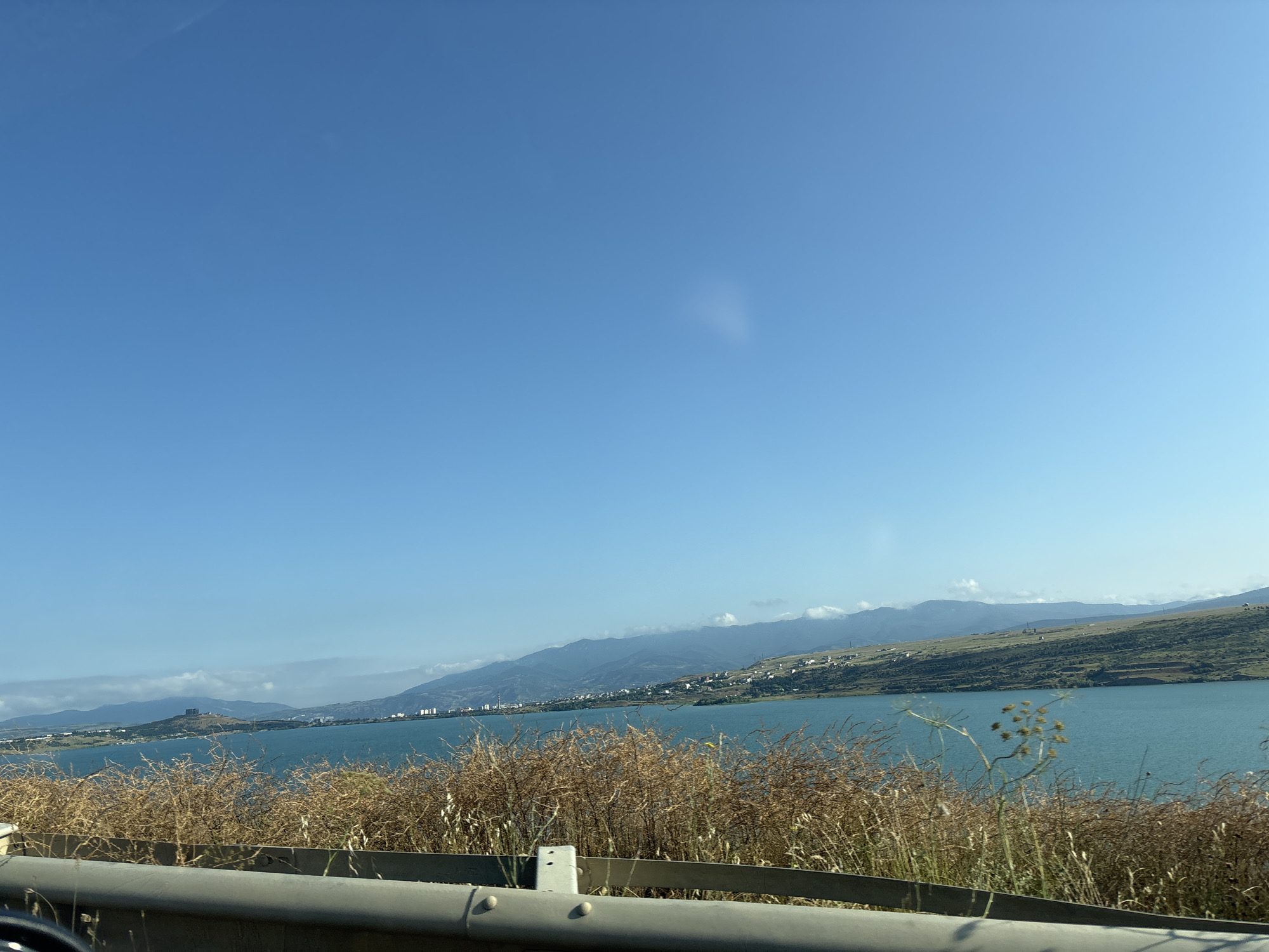 The image captures a scenic view of the Tbilisi Sea, a reservoir located northeast of Tbilisi, Georgia. The water is a tranquil turquoise, reflecting the clear blue sky. On the far bank, a range of rolling hills stretches across the horizon, dotted with scattered buildings and a prominent structure on a hilltop, likely a historical monument. The foreground is dominated by a dense growth of dry, golden-brown grasses and reeds, suggesting a warm or dry season. A gray metal guardrail runs along the bottom of the frame, indicating the viewpoint is from a road or elevated path overlooking the reservoir. The overall impression is one of a vast natural landscape meeting a developed urban fringe under a bright, cloudless sky.