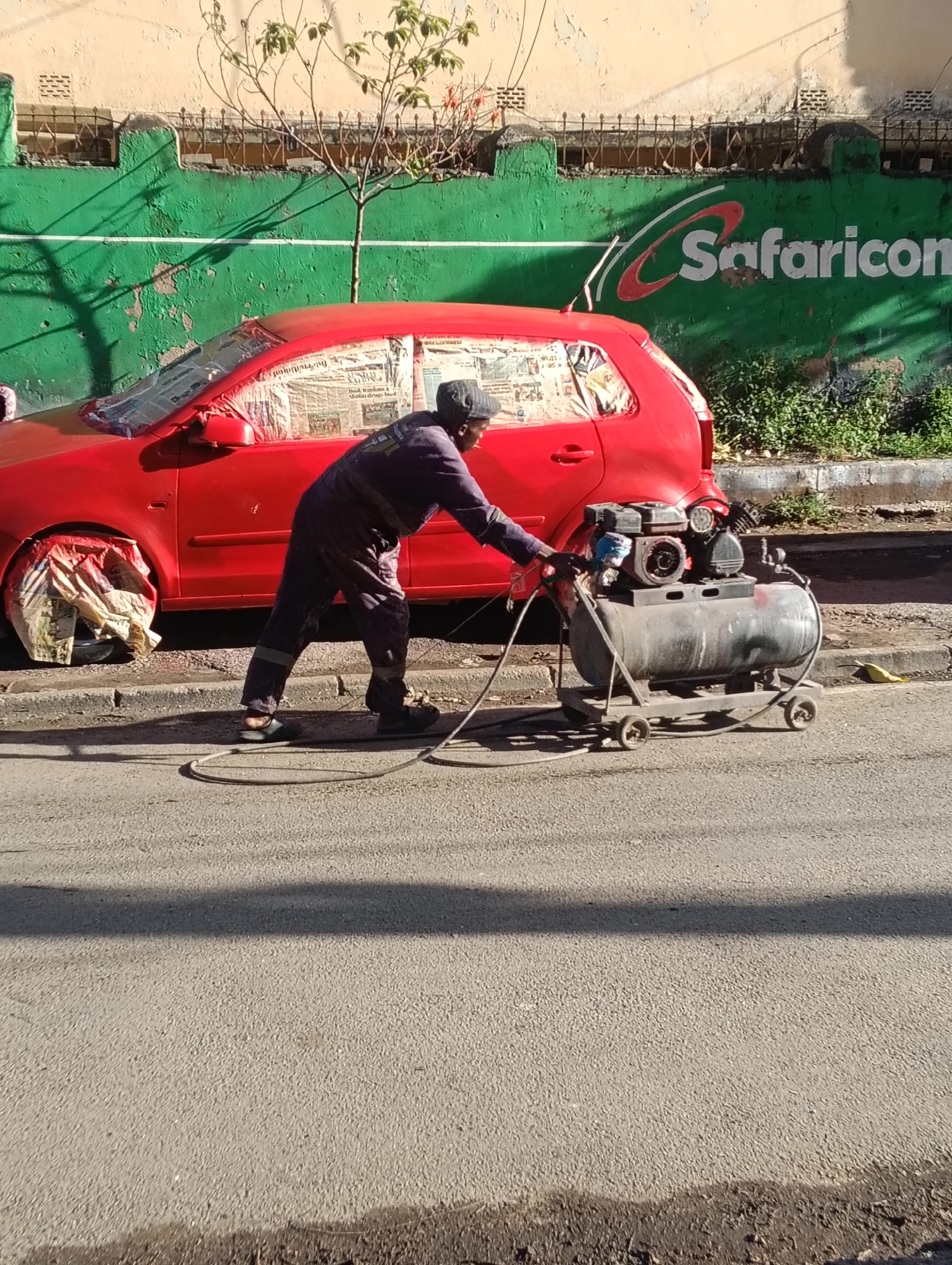 This vertical photograph captures an outdoor urban scene in Nairobi, Kenya, under bright daylight. The main subject is an individual, dressed in dark work overalls and a cap, bending over while operating equipment. They are holding a hose connected to a robust, portable air compressor mounted on a wheeled cart, suggesting involvement in spray painting.
Beside the person is a red hatchback car, prominently featured, with its windows, lights, and parts of its wheels carefully masked with newspaper and tape, indicating it is either being prepared for or is in the process of being painted.
The background consists of a green wall with peeling paint, above which a lighter-colored building or wall is visible, topped with a metal fence. A slender, young tree stands in front of the wall. On the green wall, the distinctive "Safaricom" logo is clearly visible, identifying the location as Kenya. The foreground shows an asphalt road and a curb, with a strong, horizontal shadow cast across the road, indicative of morning or late afternoon sun. Some dirt and sparse greenery are visible along the roadside.
Visible text includes the "Safaricom" logo on the wall. Newspaper fragments used for masking on the car's windows show partial text such as "DAILY NATION," "Local leaders condemn," and "bust," referencing current events. The image portrays a moment of everyday labor in a bustling street environment.