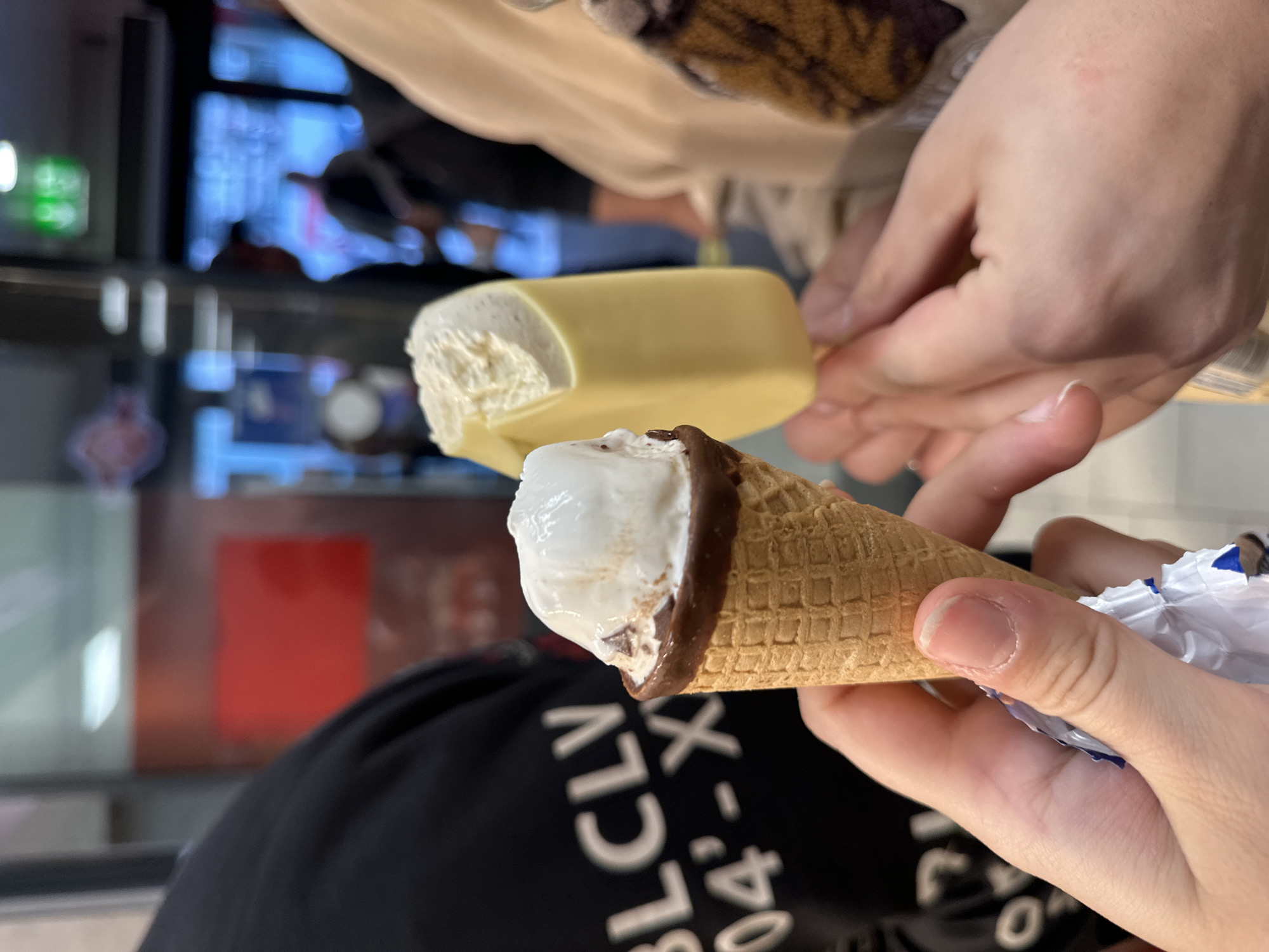A low-angle, close-up shot shows two ice creams being held. In the foreground, a person holds a cone ice cream with white ice cream and a chocolate-covered rim. The ice cream is partially unwrapped. Behind it, another person holds a partially eaten ice cream bar with a light-yellow coating. The blurred background suggests an indoor commercial space, potentially a shop or food court. A partially obscured black t-shirt with white text is visible in the foreground. The location, Varna, Bulgaria, cannot be determined from visual elements alone.