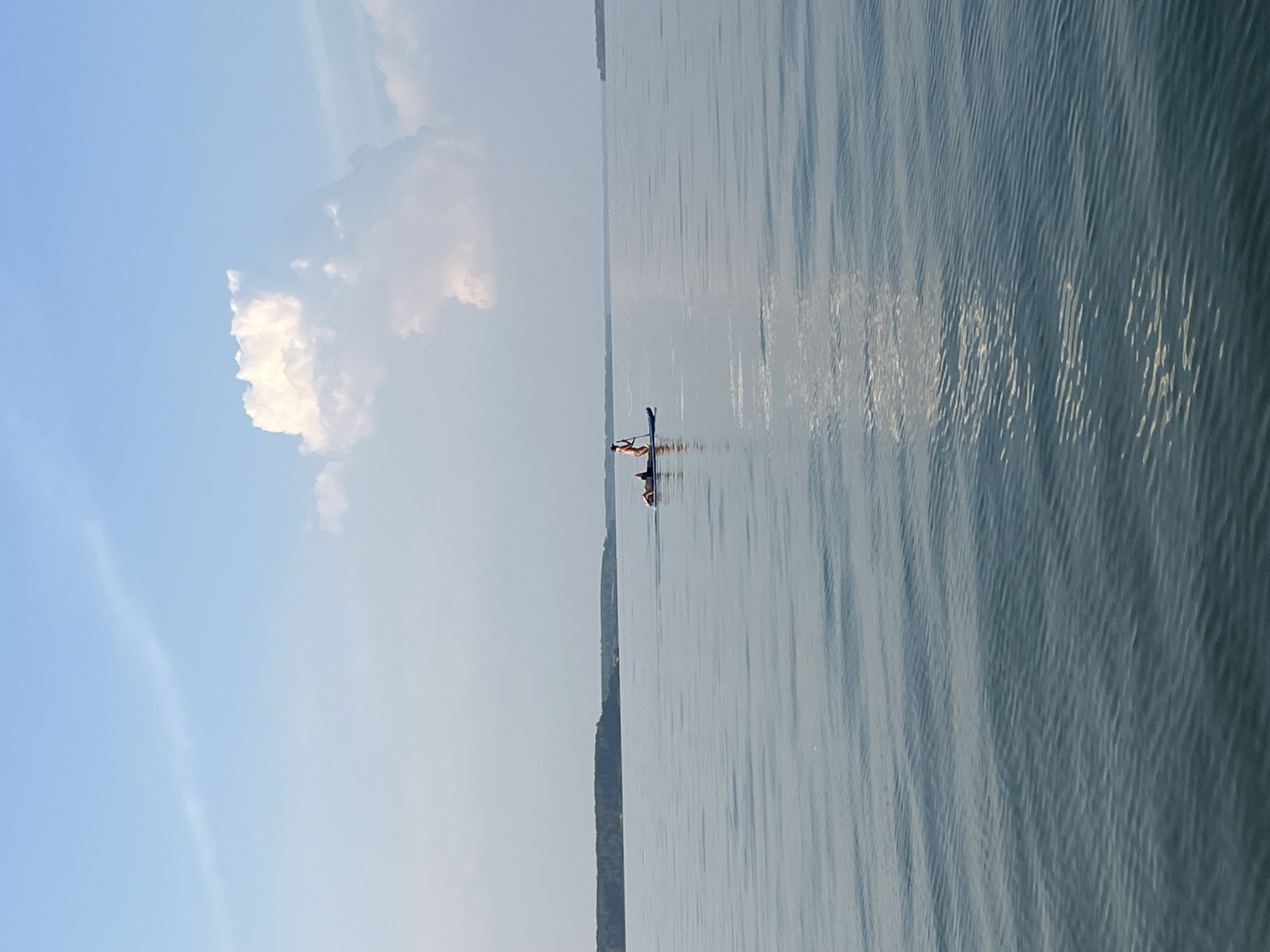 Two individuals are engaged in paddleboarding on a calm body of water. One person is standing on a paddleboard and holding a paddle, appearing to propel themselves forward. A second individual is lying down on the same paddleboard. The paddleboard is blue. The individuals are positioned in the middle of the frame, with a distant, tree-lined shore visible across the water. The sky above is a clear blue with a large, fluffy white cloud. The overall scene suggests a serene outdoor recreational activity.