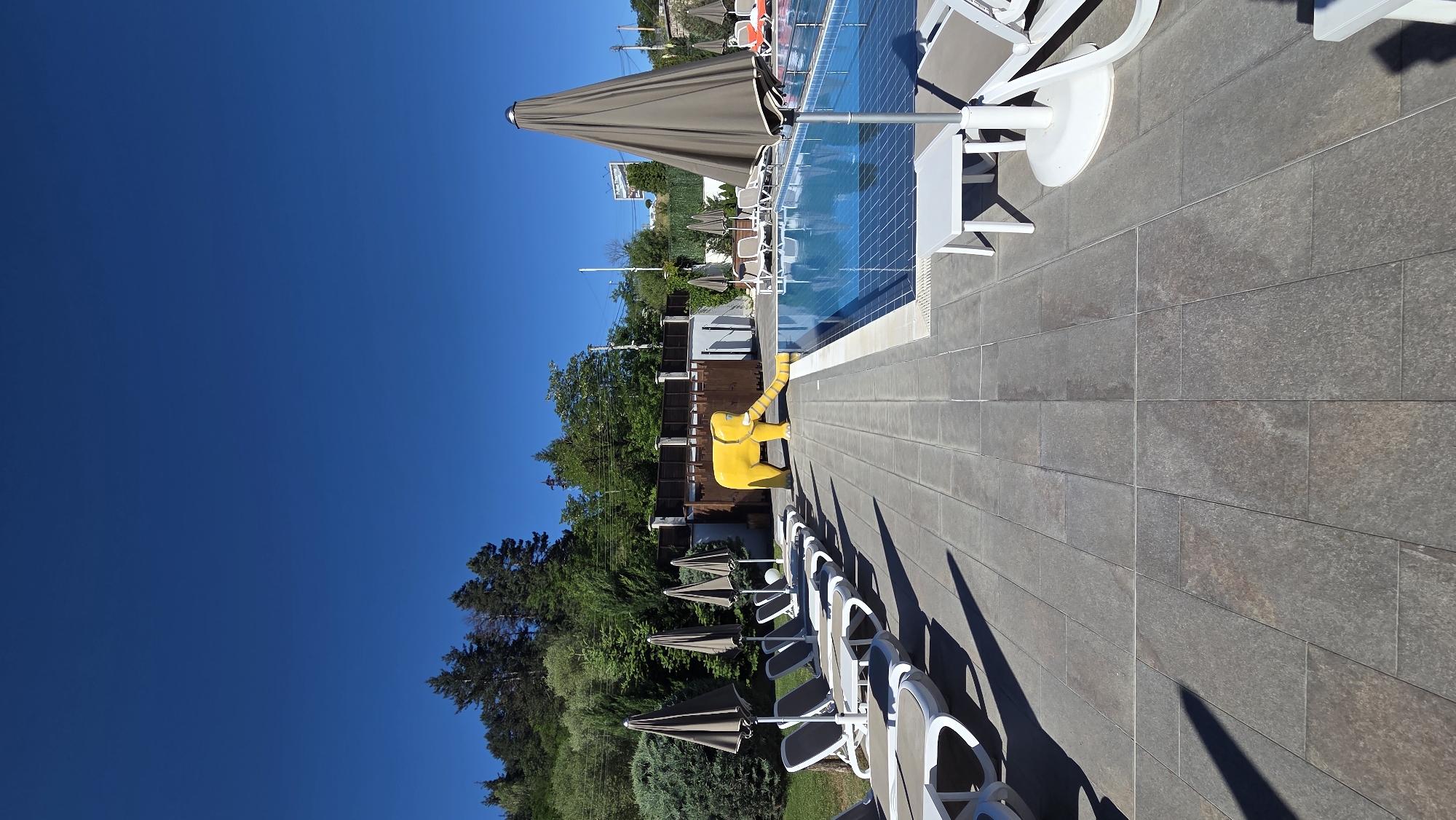The image depicts an outdoor swimming pool area on a sunny day. The pool is rectangular with bright blue water and is surrounded by a tiled patio. On the left side of the patio, several lounge chairs with gray cushions are arranged in rows, partially shaded by closed umbrellas. A small white table is placed between some of the lounge chairs.
To the right of the lounge chairs and closer to the pool, a yellow inflatable elephant slide is positioned, with its trunk extending into the pool. Behind the slide, there is a wooden fence and some buildings. Further to the right, more lounge chairs and umbrellas are visible. The background features lush green trees and a clear blue sky. The overall scene suggests a recreational area, possibly a resort or a public swimming facility.