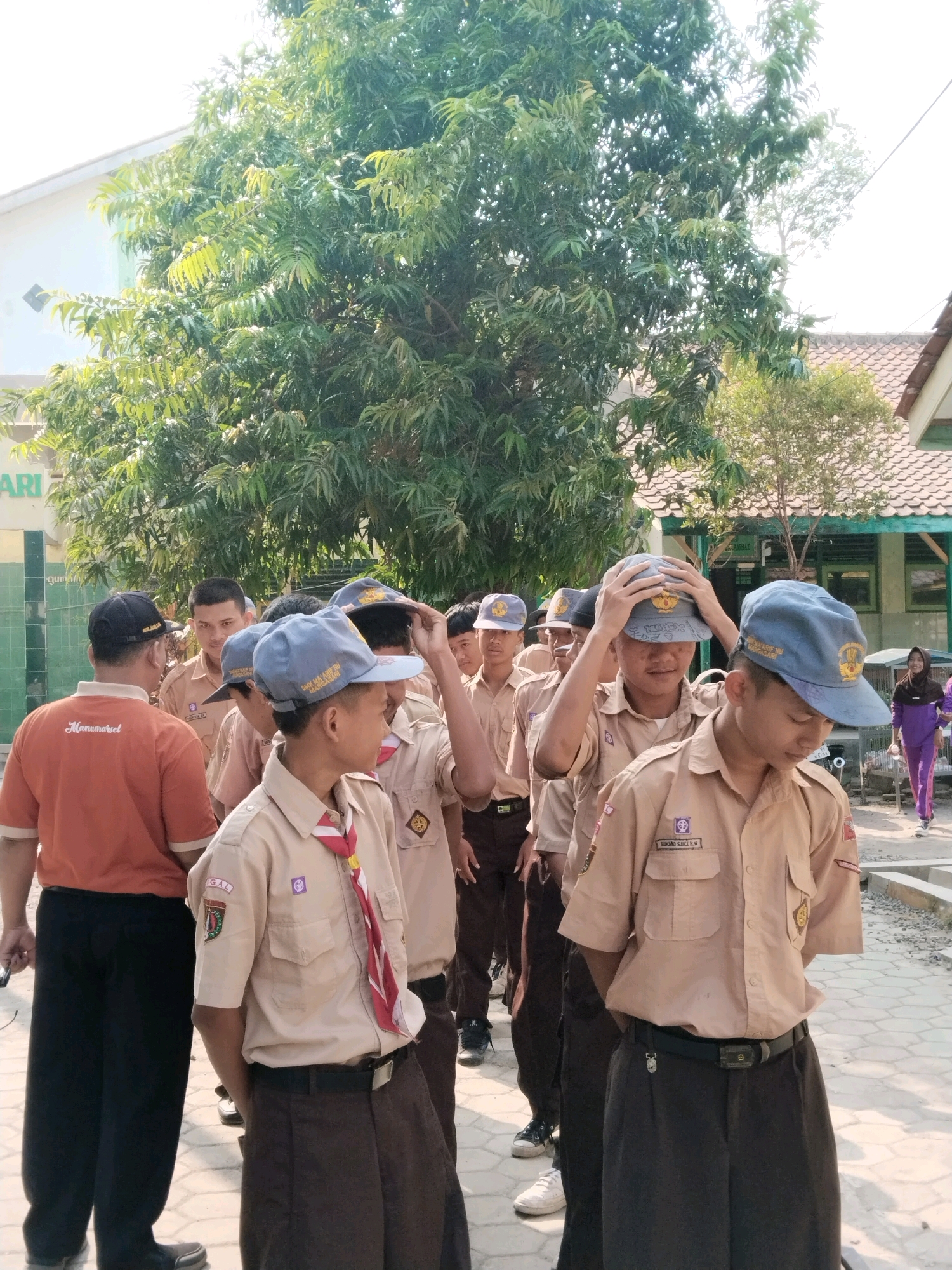 The image captures a group of male students in uniform standing outdoors. The students are wearing beige shirts with emblems and badges on the sleeves, brown pants, and hats. Some of them have red and white striped neckties. The setting appears to be a schoolyard in Pakulaut, Indonesia, with a building and a large tree in the background. The students seem to be lined up or gathered, possibly for an activity or assembly. The lighting suggests it is daytime.