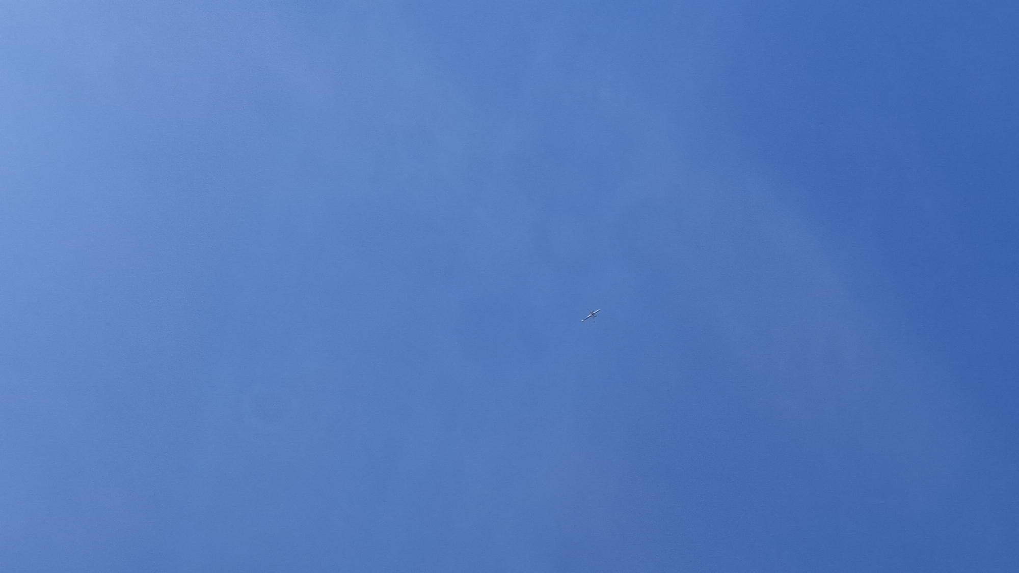 The image is a vertical, low-angle shot of a clear, bright blue sky. A single, small, white airplane is visible in the center of the frame, flying from left to right. The airplane appears to be at a high altitude. The sky is uniform in color, with no clouds or other atmospheric disturbances. The image was taken in the city of Smolevichi, Belarus.