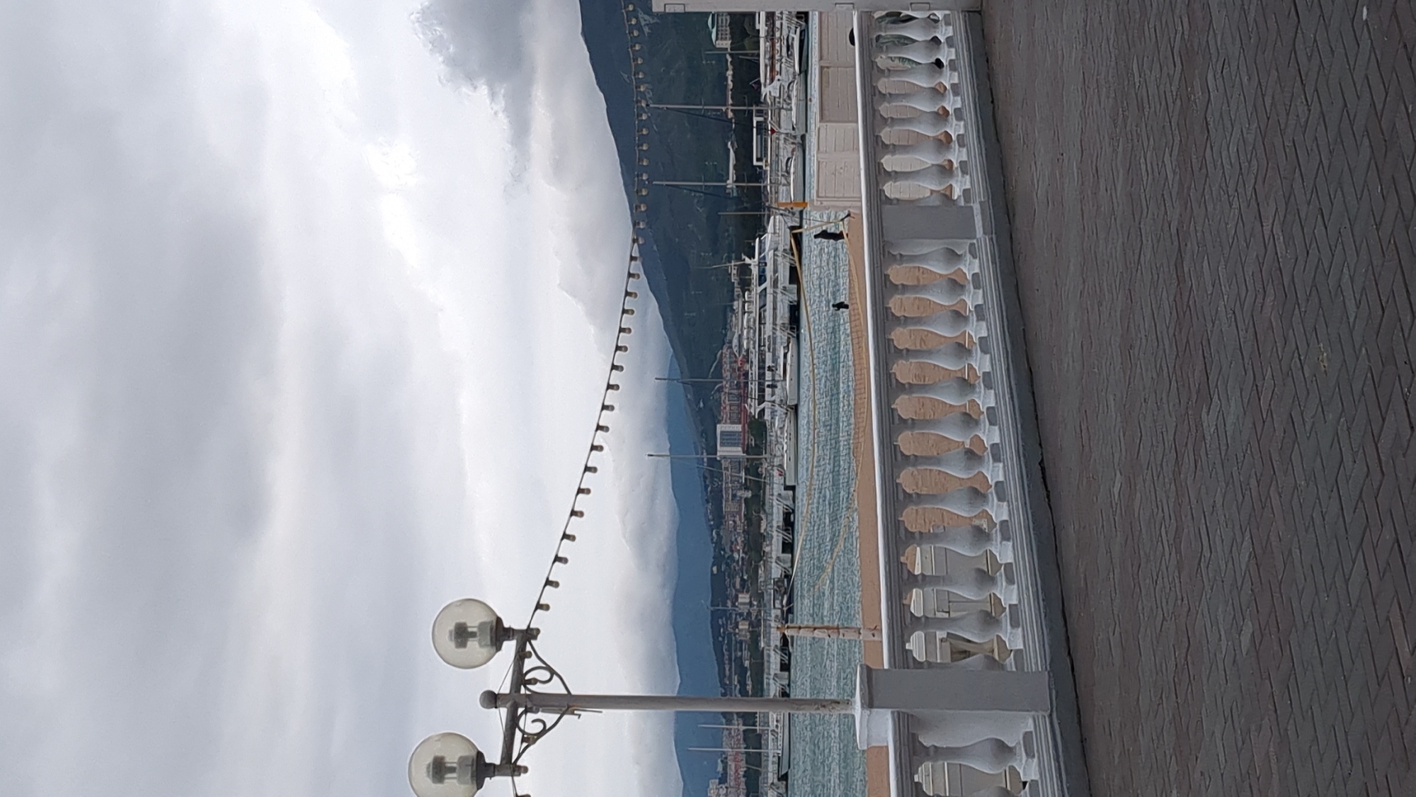 A waterfront scene in Gelendzhik, Russia, showcases a promenade lined with a decorative white balustrade and brick pavement. In the distance, a mountain range rises amidst a cloudy sky. A row of boats and yachts are moored in the harbor. A vintage street lamp stands nearby. The lighting suggests an overcast day.