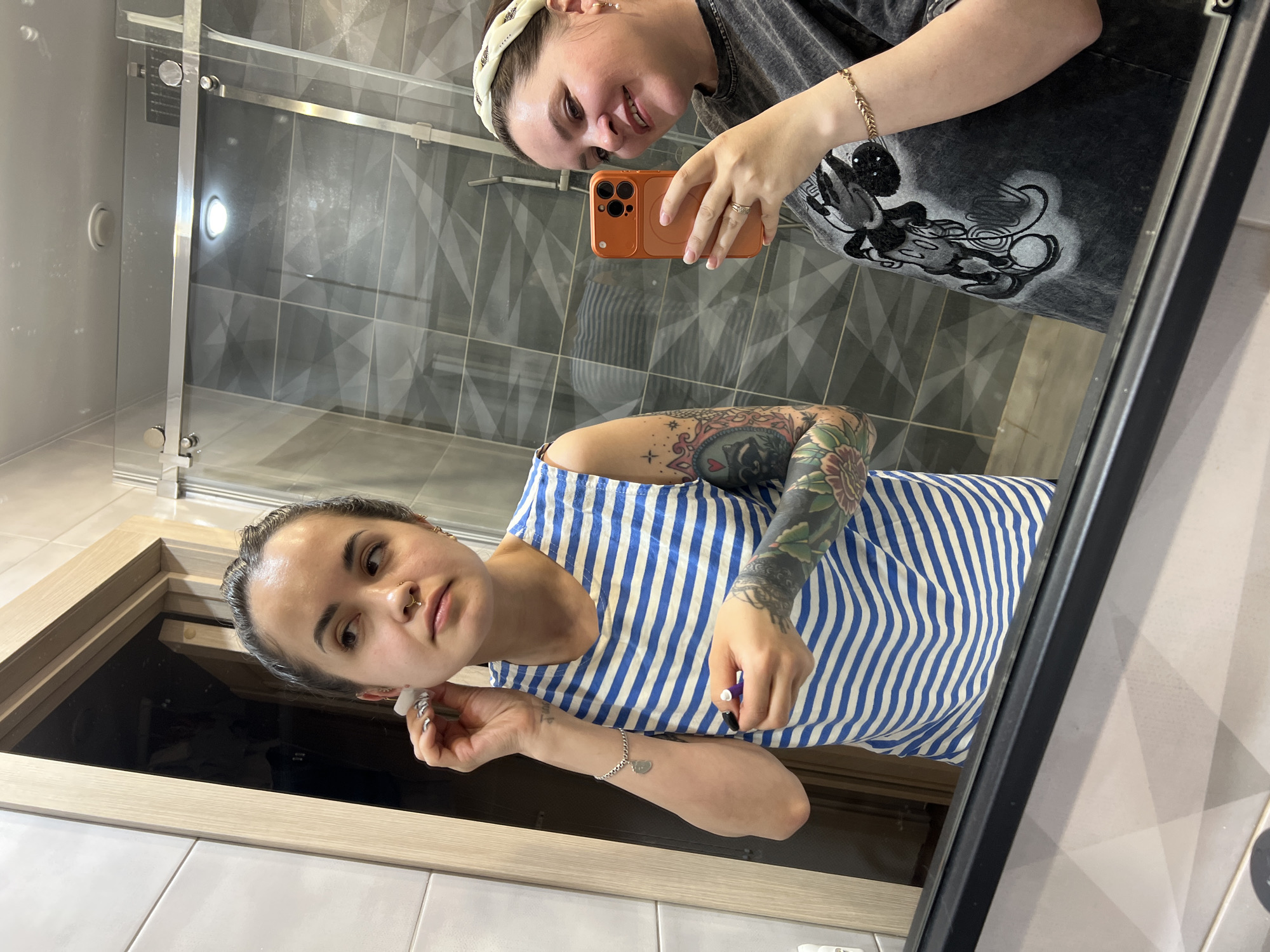 A woman with arm tattoos and a striped shirt is visible in a mirror selfie. In the reflection, another woman is holding an orange phone, presumably taking the photo. The setting appears to be a bathroom, suggested by the presence of tiles and a glass shower screen. No information is provided that indicates the location of the bathroom is in the city of Noviy, Russia.