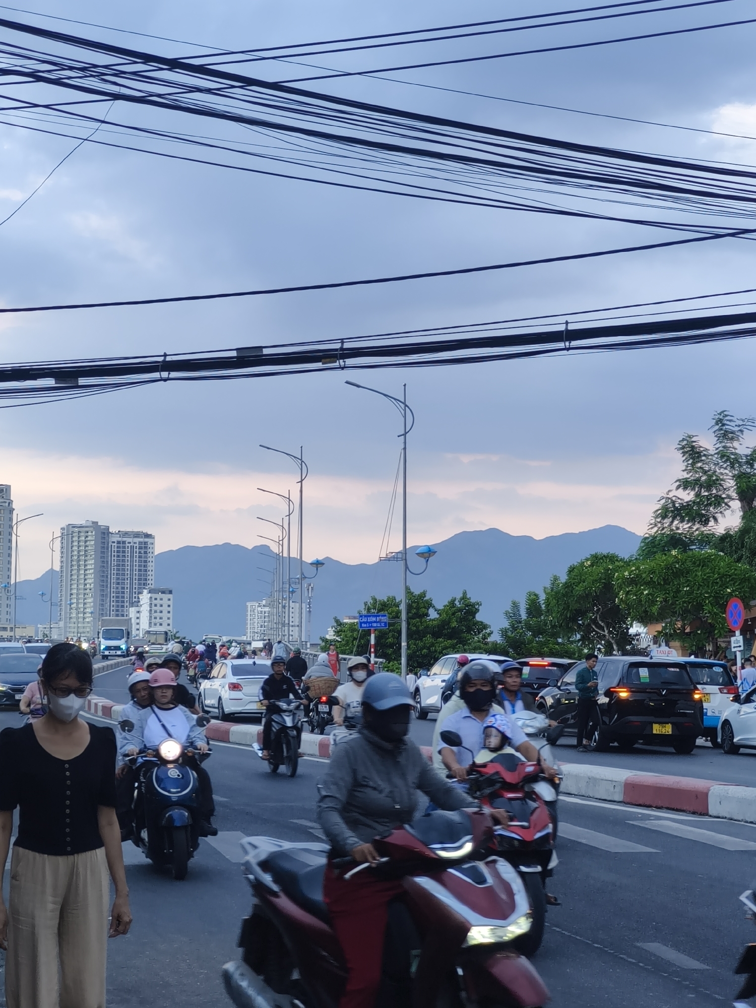 A street scene in Bac Nha Trang Ward, Vietnam, is captured from a low angle looking towards a road filled with traffic, including motorcycles, cars, and a truck. The sky is overcast with a pastel hue, suggesting either dawn or dusk, and is crisscrossed by numerous overhead electrical wires. In the background, several modern high-rise buildings stand against a backdrop of hazy mountains.
On the road, a woman wearing glasses and a white face mask, a black short-sleeved top, and beige pants walks towards the viewer in the foreground, slightly to the left. She is the most prominent individual. To her right and further down the road, multiple motorcycles are visible, some carrying two or more passengers. One motorcycle in the mid-ground has a rider wearing a blue helmet and a passenger with a pink helmet. A blue and white scooter is also present. Several cars are also in traffic, including a black SUV with a yellow license plate that reads "79H 116.24", a white sedan, and a white truck in the distance. Traffic lights and street lamps line the road. Trees and foliage are visible on the right side of the image, and a street sign with Vietnamese text is mounted on a pole. The road has a white and red striped curb.