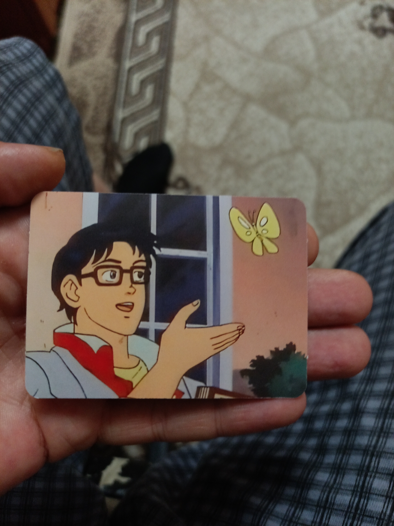 A person is holding a rectangular card depicting an anime-style illustration. The illustration features a young man with black hair and glasses, wearing a red and white collared shirt and a light blue jacket. He has an open-mouthed expression of surprise or confusion. A yellow butterfly with white spots is flying near his outstretched hand. The background shows a window with blue panes and a peach-colored sky. The card is held in a hand, with the person's thumb and fingers visible. The background appears to be a bed with a textured blanket and a person wearing plaid clothing. The image was taken in the city of Nyagan, Russia.