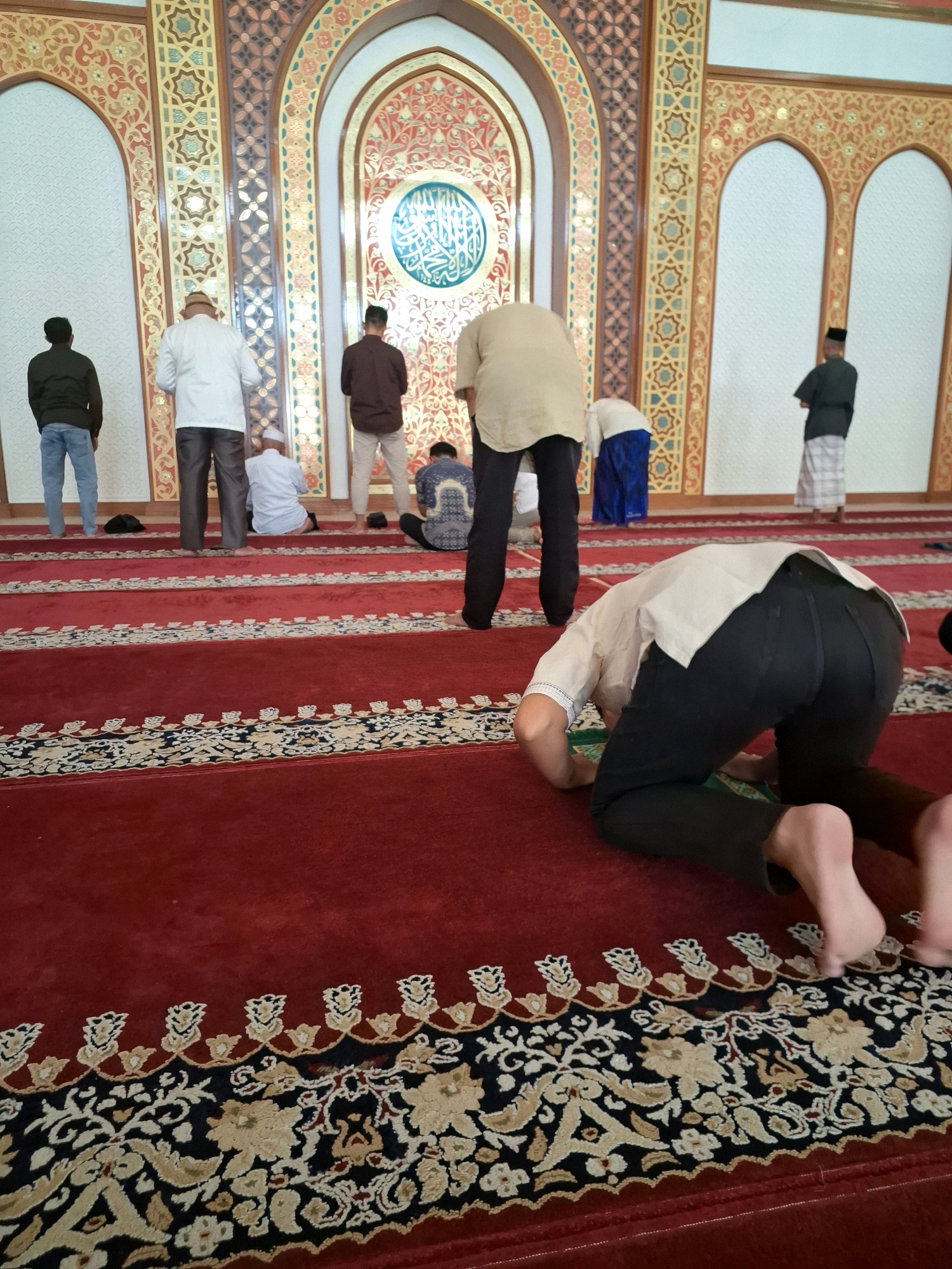 The image depicts a group of men engaged in prayer inside a mosque. The mosque features ornate walls with geometric patterns and Arabic calligraphy, indicative of Islamic architecture. The floor is covered with a red carpet with intricate designs. Several men are in various stages of prayer, including standing and prostrating. One man in the foreground is in a prostrating position, with his head bowed low. Other men are seen standing or kneeling in prayer further back. The men are dressed in traditional or casual attire. The setting appears to be a place of worship within the city of Gorontalo, Indonesia, as suggested by the context.