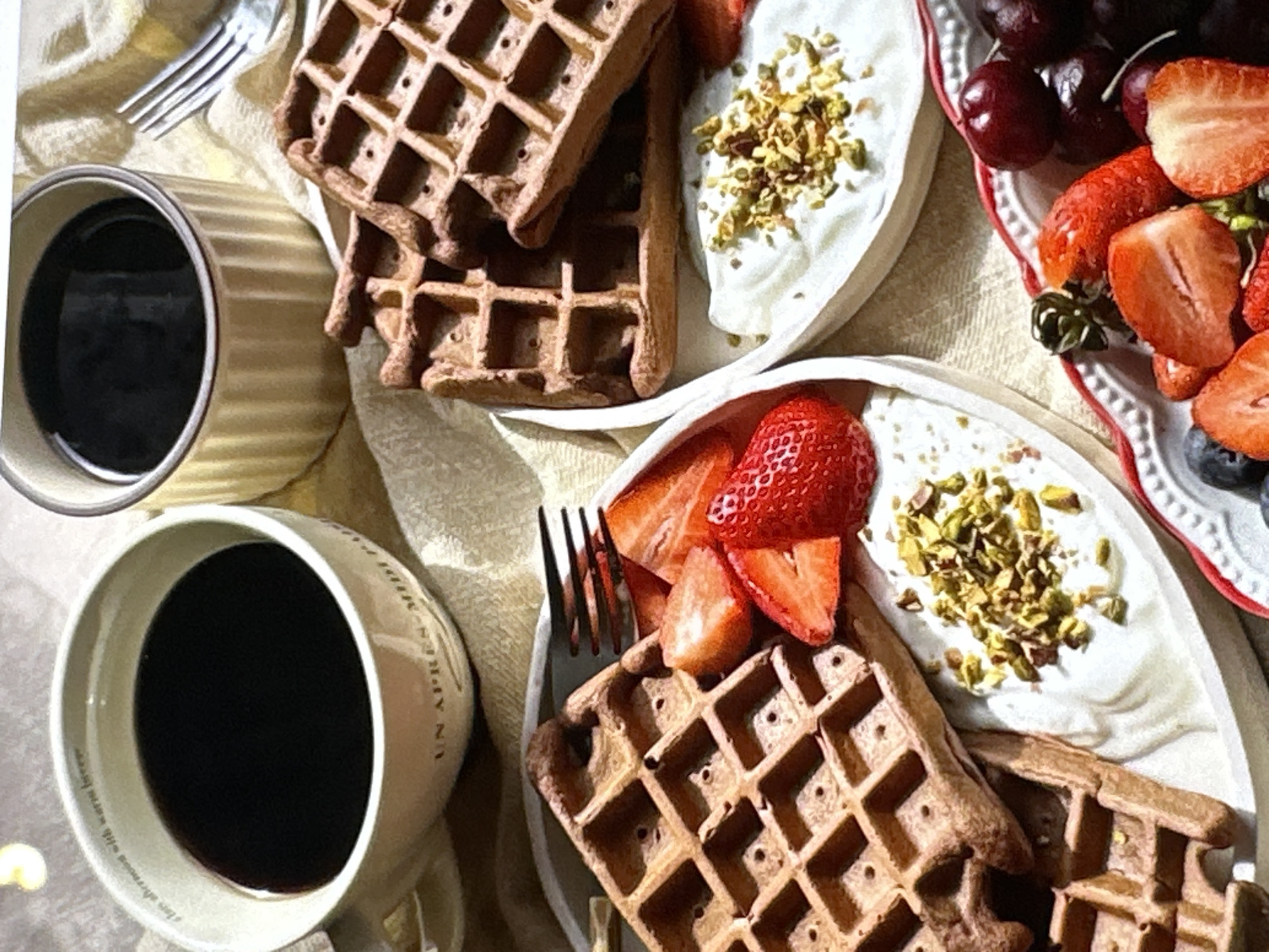 The image depicts a breakfast scene with two plates of waffles, a plate of berries, and two mugs of coffee. The waffles are topped with what appears to be yogurt and chopped pistachios, accompanied by sliced strawberries on one plate. The berry plate contains strawberries, cherries, and blueberries. A fork is placed on the table near each plate of waffles, and the mugs are filled with dark coffee. The scene is set on a light-colored cloth. The specific location, Yerevan, Armenia, cannot be determined from the image alone.