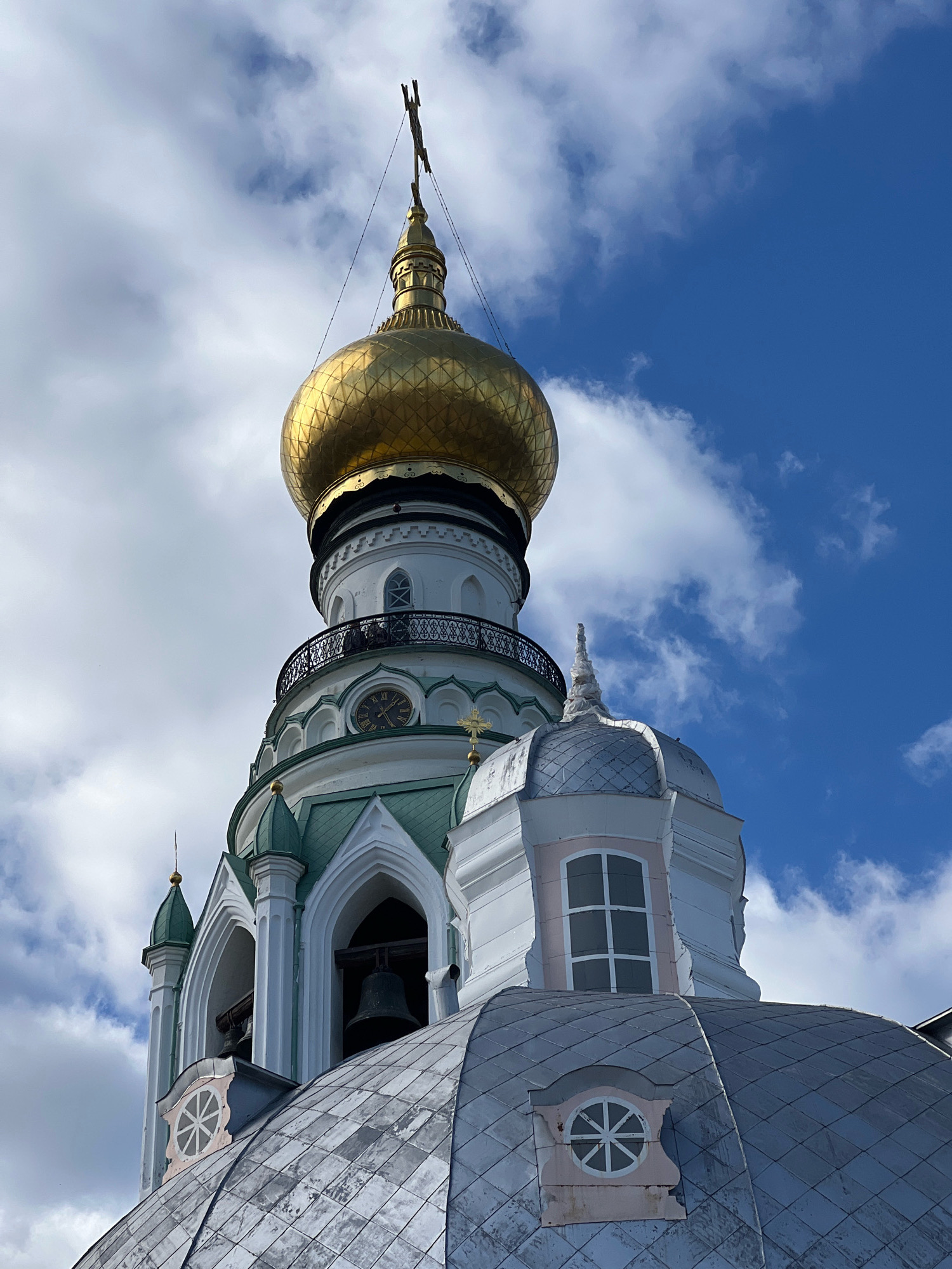 A low-angle, close-up, full-shot photograph shows a gold-domed Russian Orthodox church bell tower against a bright blue sky with white clouds. The bell tower is white with green trim and has a clock face visible. Below the gold dome, a bell hangs in an arched opening. A silver-tiled dome is visible in the foreground. The image appears to be taken in daylight.