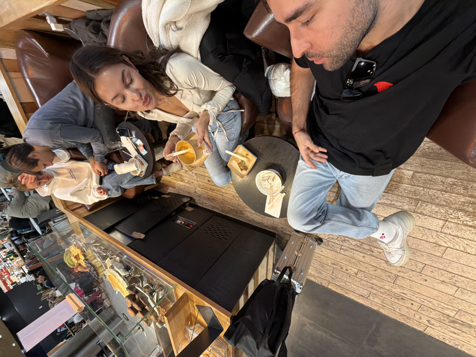The scene depicts four individuals seated in a cafe or food court within Colombier-Saugnieu, France, likely at Lyon-Saint Exupéry Airport.
In the foreground, a male with dark hair and a beard, wearing a black t-shirt with a red heart emblem and light blue jeans, is seated in a brown upholstered armchair. He looks towards the left with his eyes partially closed, his right hand resting on his thigh. White socks with a red graphic and white sneakers are visible. Sunglasses are tucked into his shirt collar. To his immediate left, a black backpack rests on a silver-colored rolling suitcase with an extended handle.
In the mid-ground, a female with long dark hair, wearing a light-colored long-sleeved top and light blue jeans, is seated in a brown upholstered chair opposite a dark circular table. She holds a cardboard bowl containing an orange-colored food item with a wooden spoon, bringing the spoon towards her mouth. Her lips are slightly puckered. A white jacket is draped over the back of her chair. On the table, an empty cardboard bowl with a wooden utensil and a crumpled napkin are present.
In the background, two individuals occupy brown upholstered seating. A female with light brown hair and glasses, wearing a white hooded sweatshirt and blue jeans, is seated, looking down at a mobile phone she holds. Beside her, a male with short dark hair, wearing a grey long-sleeved shirt and dark pants, leans towards her, also looking down, potentially at the same device. A white paper cup, a mobile phone, and some banknotes are on the dark circular table in front of them.
To the left of the image, a food service counter features a glass display case showcasing various baked goods, including rows of muffins, a large tart, and fresh fruit such as bananas. The counter base is dark-colored. The floor throughout the seating area is composed of light brown wooden planks, transitioning to a darker, smooth surface near the luggage. Wooden paneling lines the wall behind the seated individuals, and the area is illuminated by artificial lighting.