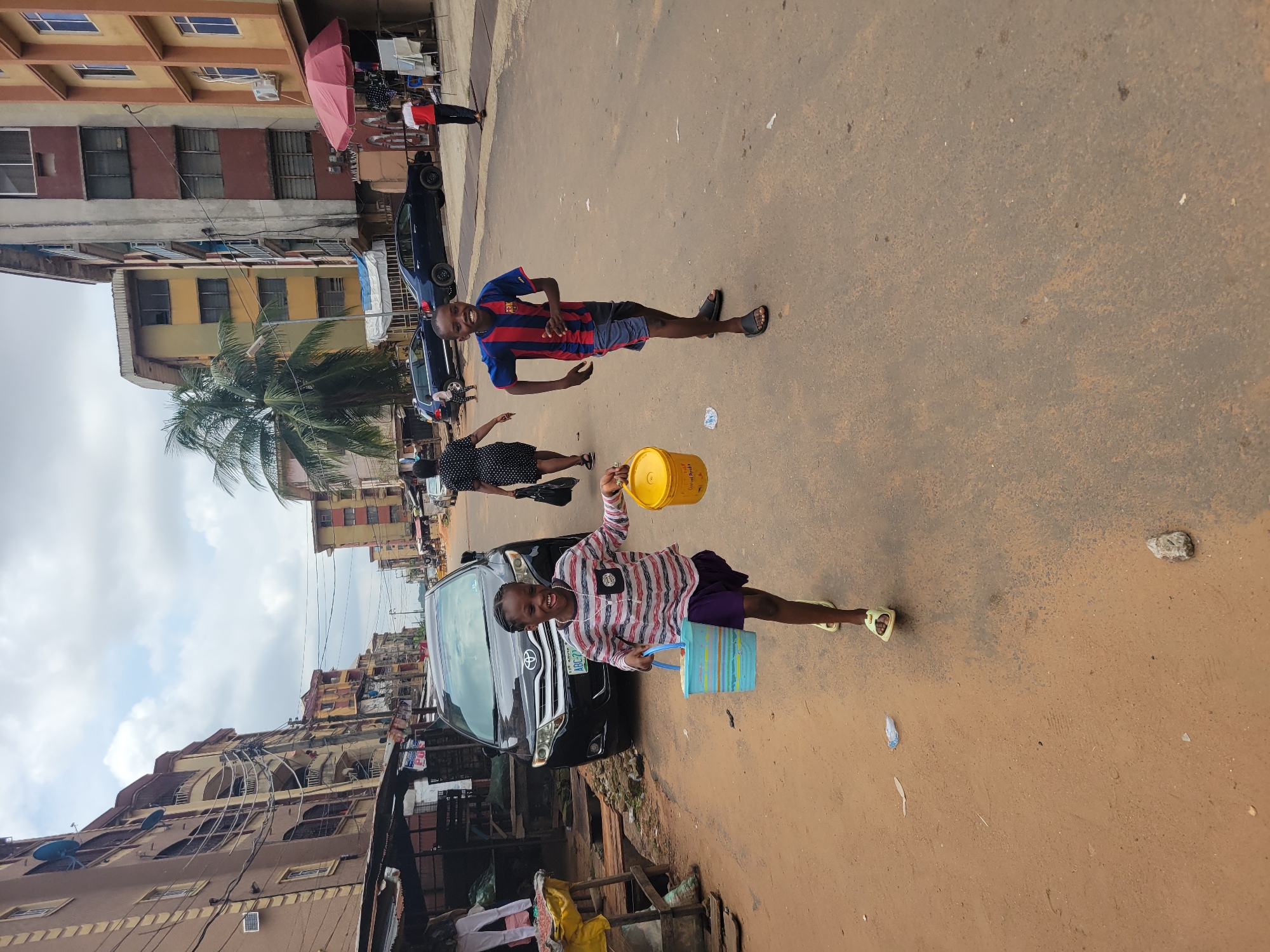 In Onitsha, Nigeria, two children are captured running down a dirt and asphalt road. The girl in the foreground, wearing a striped shirt and purple skirt, grins as she carries a blue bucket in her left hand and a yellow one in her right. Behind her, a boy in a blue and red striped soccer jersey and grey shorts jogs with a joyful expression. Their surroundings are typical of a busy urban area; the road is lined with multi-story buildings, some painted in earthy tones, others in faded yellows and reds, with visible electrical wiring and satellite dishes. A dark SUV is parked on the side of the road, and a woman in a black and white polka dot dress walks in the background. A red umbrella provides shade over a market stall, suggesting commercial activity. The sky above is overcast with scattered clouds, indicating a warm, humid climate. The scene depicts a moment of childhood energy amidst the everyday life of Onitsha.