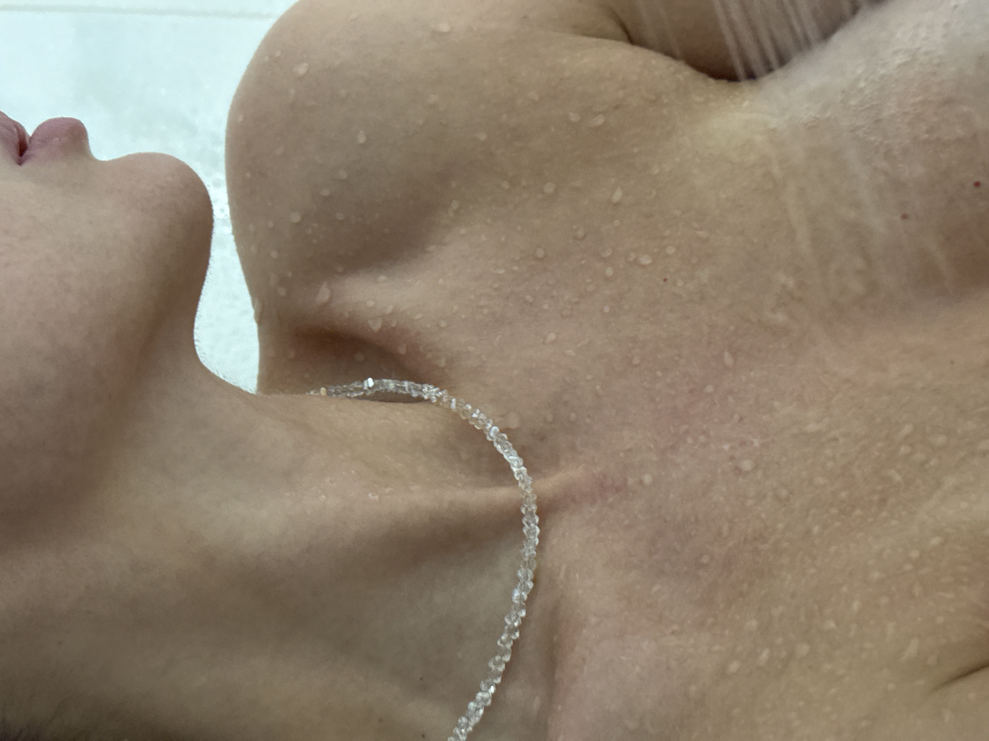 The image shows a person's neck, chest, and part of their face in what appears to be a shower. The person is fair-skinned and wet, with water droplets visible on their skin. They are wearing a delicate, beaded necklace. The background is a blurry white, suggesting a bathroom or shower stall. The location is unknown and there is no indication the image was taken in Chaikovsky, Russia.