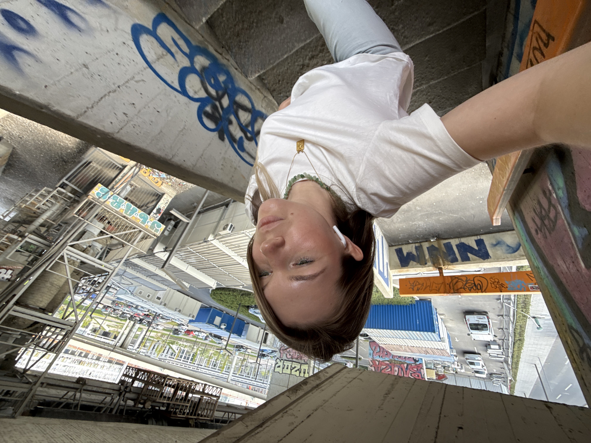 The media file, presented on its side, prominently features a young woman looking directly into the camera. She appears to be looking upwards from beneath a concrete structure. She has light skin, blue eyes, and medium-length brown hair, and is wearing a white t-shirt, light grey pants, a gold pendant necklace, a green beaded necklace, and white wireless earbuds. Her expression is neutral and calm.
The setting is an outdoor urban environment, captured during bright daylight, likely under sunny conditions. The woman is framed within a gritty concrete structure, possibly a bridge, overpass, or multi-story parking garage in Belgrade, Serbia. The concrete surfaces around her are heavily adorned with graffiti. Visible text includes "WNN" in bold blue letters on a large concrete beam. Further back, "4OZO" or a similar numerical text is spray-painted in green on a vertical concrete surface.
Beyond the immediate concrete structure, the background reveals an expansive view of ground level. This includes metal scaffolding or structural elements, various buildings, a road with several parked or moving vehicles (vans and cars), and large blue and white shipping containers. Patches of green foliage are also discernible, adding a touch of nature to the industrial cityscape. The overall scene conveys a sense of depth and a contrast between the immediate, intimate selfie and the sprawling urban backdrop.