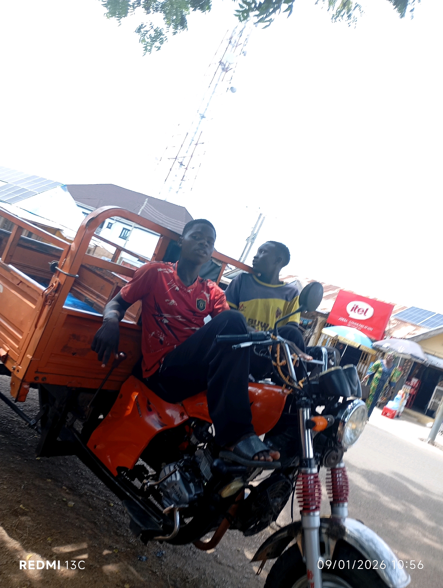 Two young men are seated on a red and orange motorized tricycle, parked on a dirt and paved road. The man in the foreground, wearing a red t-shirt with a graphic print and black pants, is sitting on the rear seat of the tricycle and looking to the left. The man behind him, wearing a yellow and gray t-shirt and black pants, is on the driver's seat, looking forward. The tricycle has a large orange cargo bed at the back. In the background, there are buildings with solar panels on their roofs, a red billboard with the "itel" logo, and other people walking. The scene appears to be in a market area in Shendam, Nigeria, suggested by the presence of stalls and goods for sale. The time stamp on the image indicates January 1, 2026, at 10:56.
