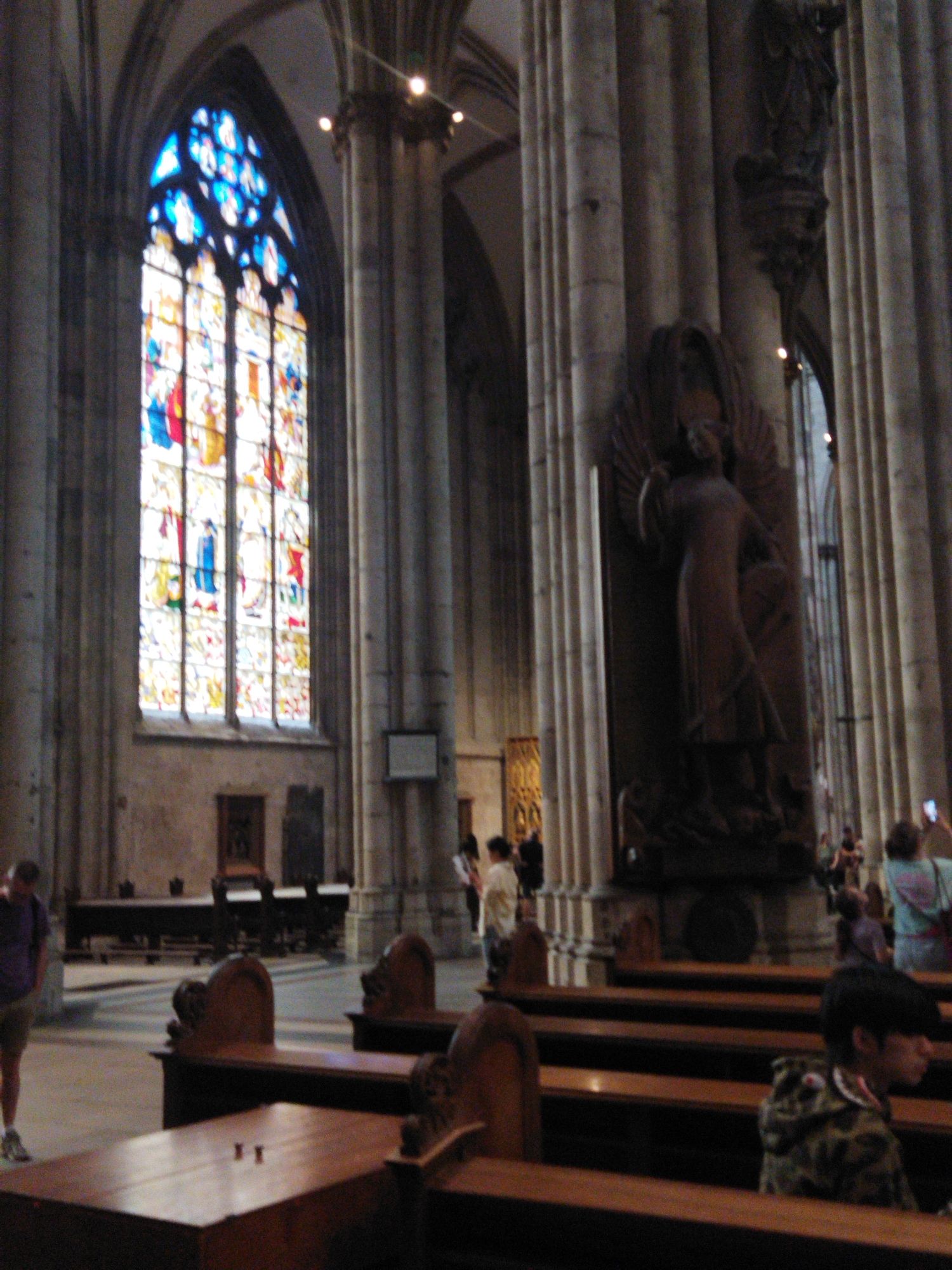 An interior shot shows a large cathedral with tall columns and a stained glass window. A man in a purple shirt and shorts stands in the foreground to the left. To the right, several people are seated on wooden pews. In the background, a large statue of an angel is visible. The scene appears to be within the Monheim am Rhein region of Germany, likely in a prominent church.